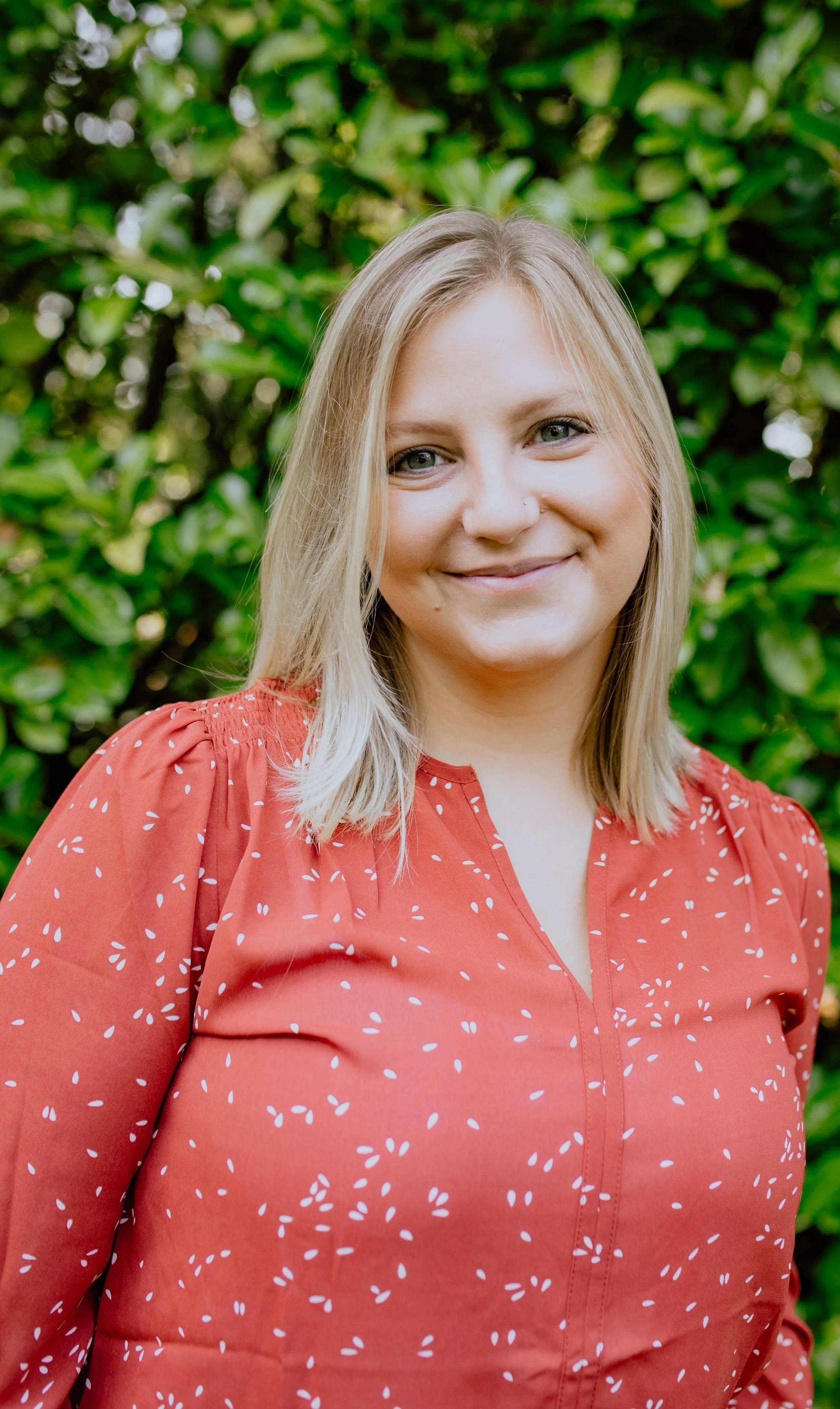 A woman with blonde hair and a nose piercing wearing a red blouse with white patterns, standing outdoors in front of green foliage, smiling at the camera. Seattle professional head shot photography