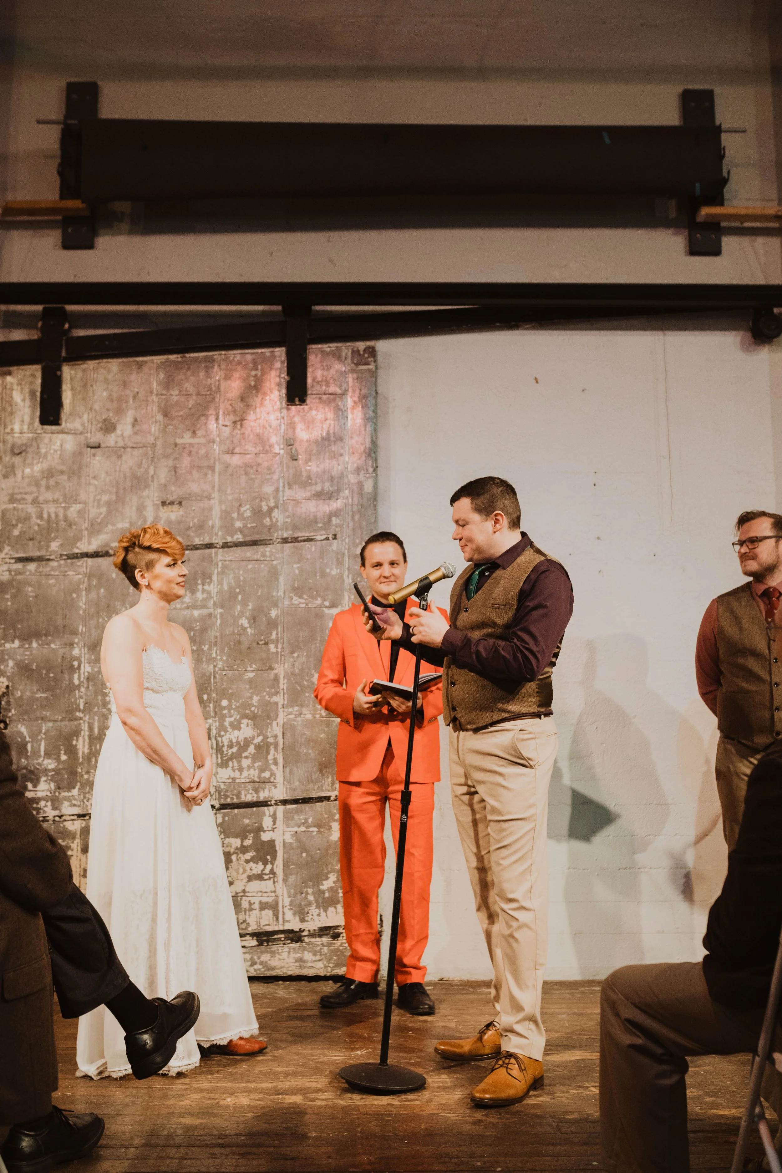 A man in a brown vest and beige pants is giving a speech or reading vows at a wedding ceremony. A woman in a white wedding dress watches him. Two other people, one in a bright orange suit and the other in a vest and tie, stand nearby on a rustic wood