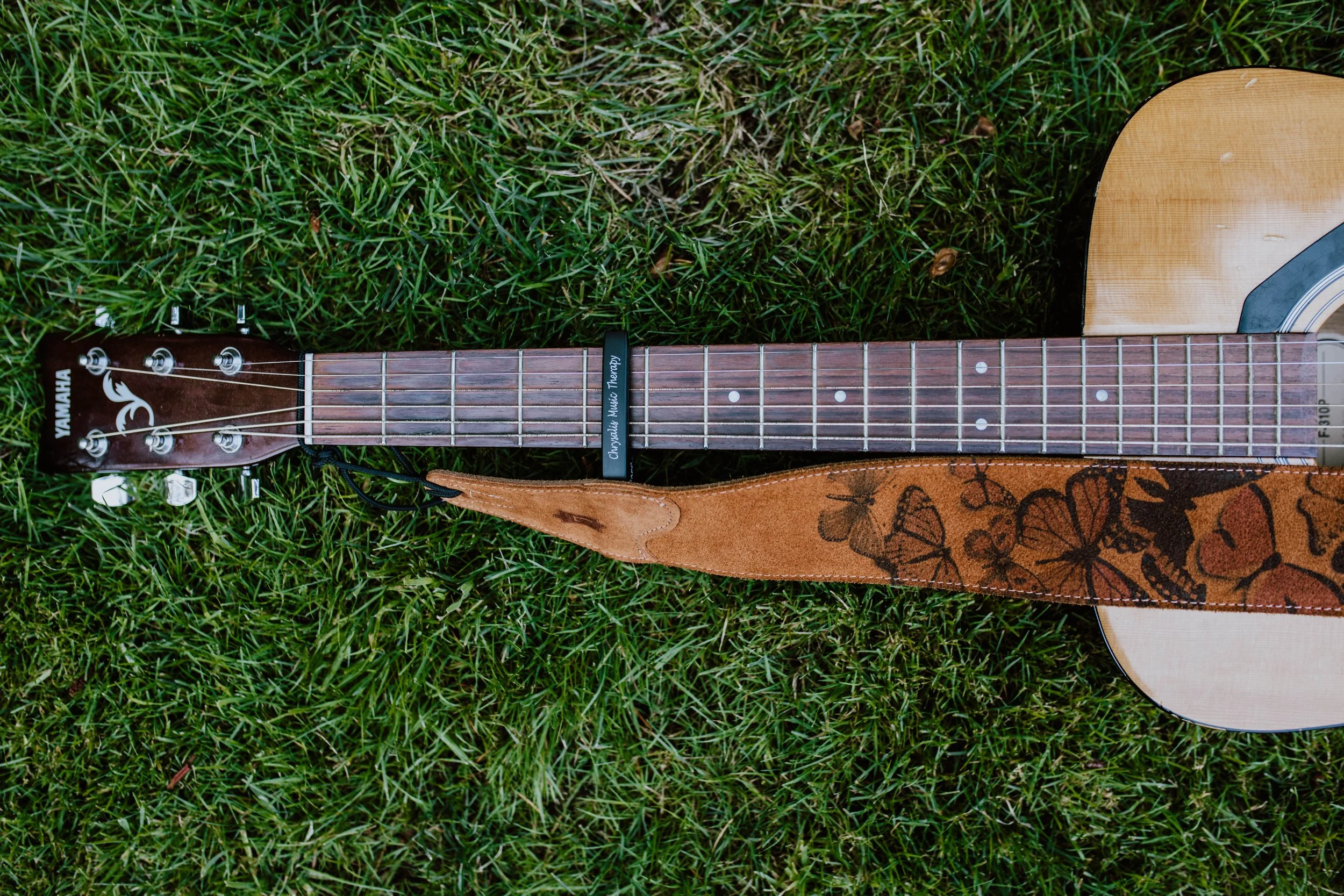 An acoustic Yamaha guitar lying on grass, with a leather strap decorated with butterflies, and a capo placed on the neck of the guitar. Seattle professional head shot photography