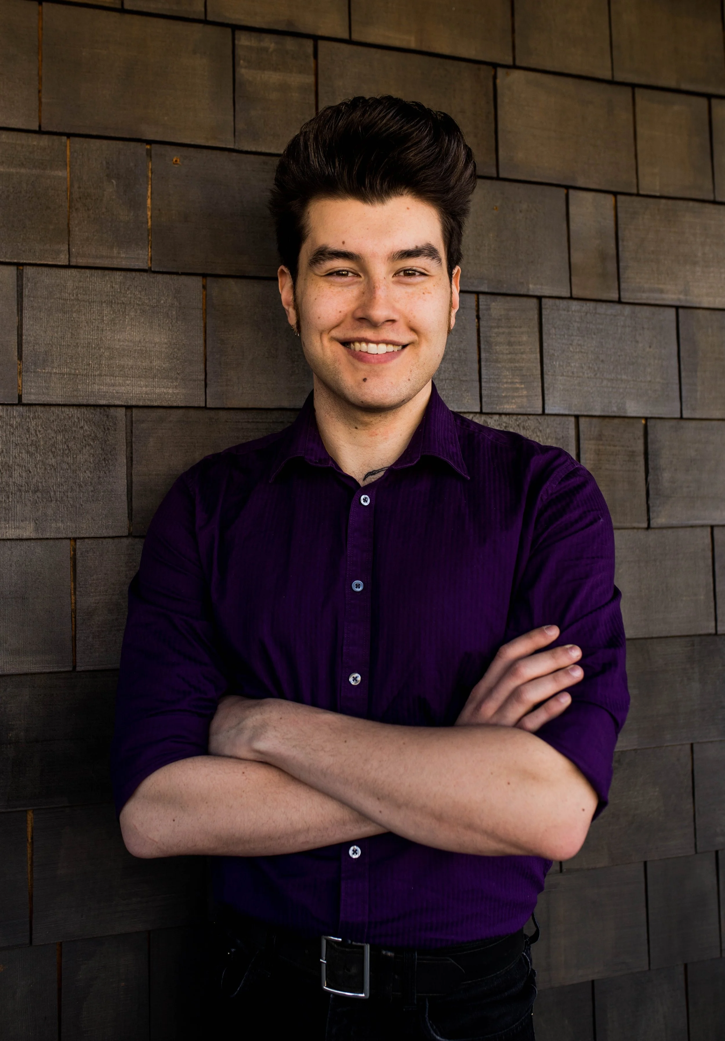 Young man with short dark hair, smiling, wearing a purple shirt, standing with arms crossed in front of a wood-paneled wall. Seattle professional head shot photography