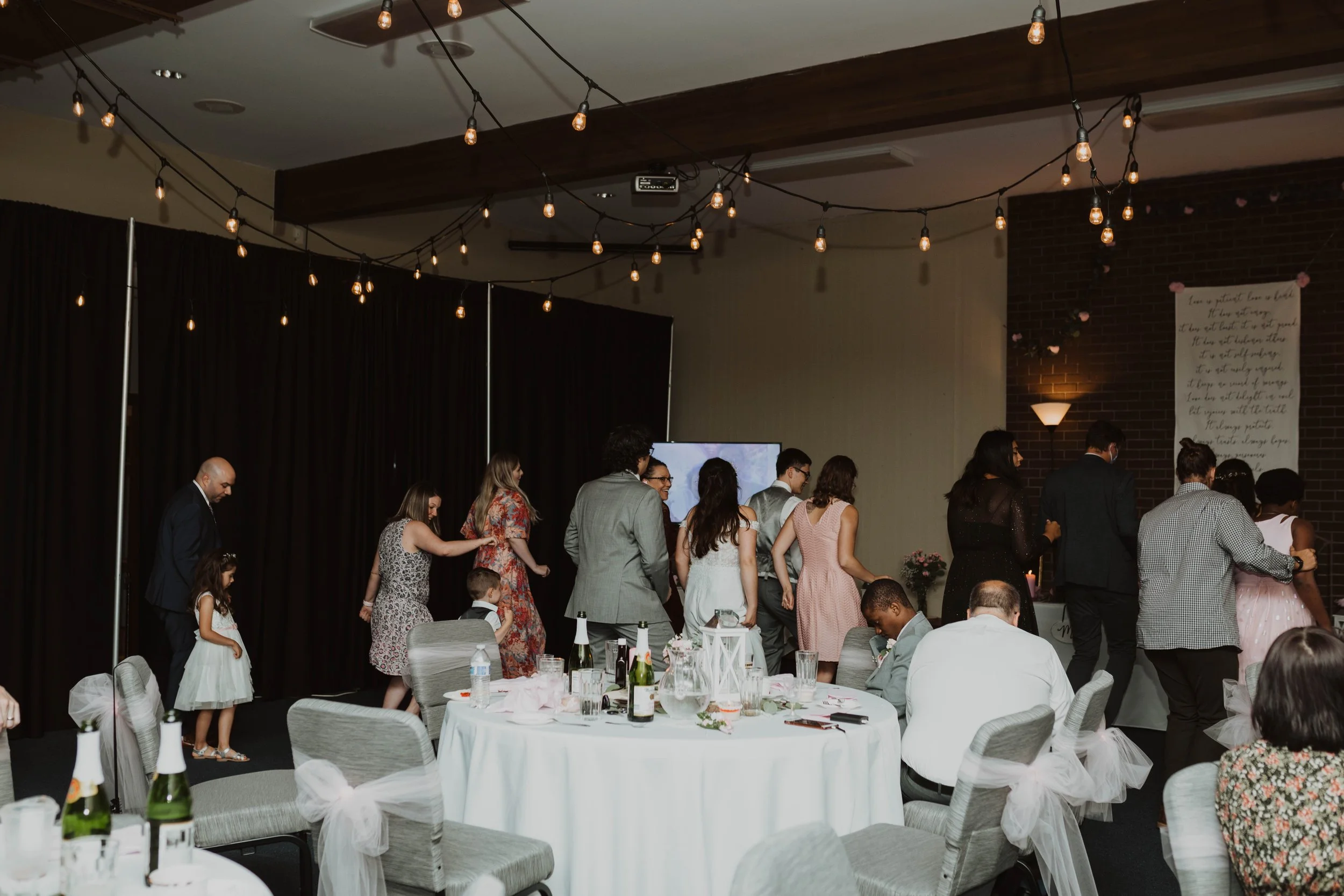 People, including children and adults, standing in a line at a wedding reception or banquet. The room has round tables with white tablecloths, decorated with bottles, glasses, and floral centerpieces. The ceiling is adorned with string lights, and th