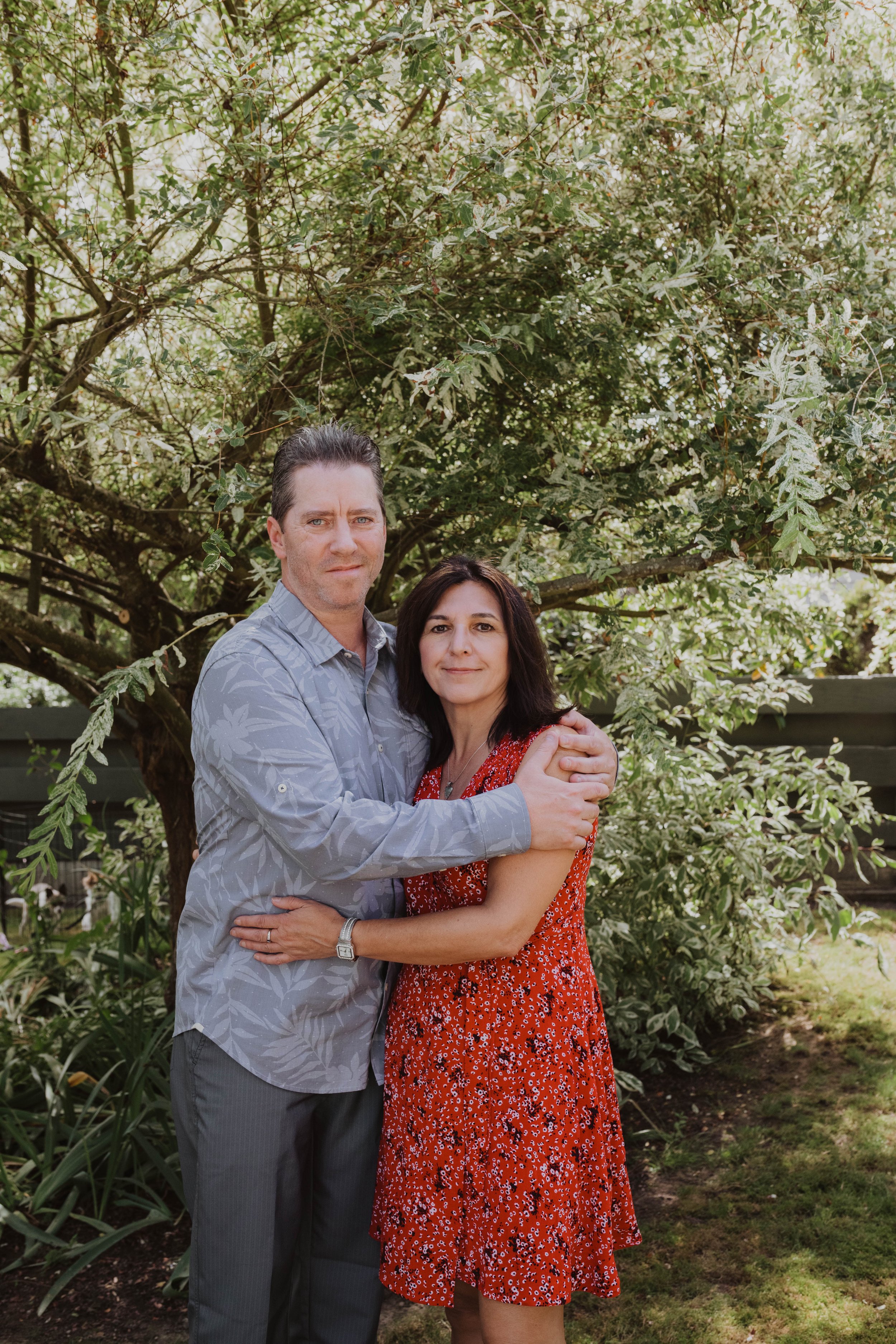 A man and woman standing together outdoors in front of a tree, embracing each other and looking at the camera. Seattle, WA wedding photography.