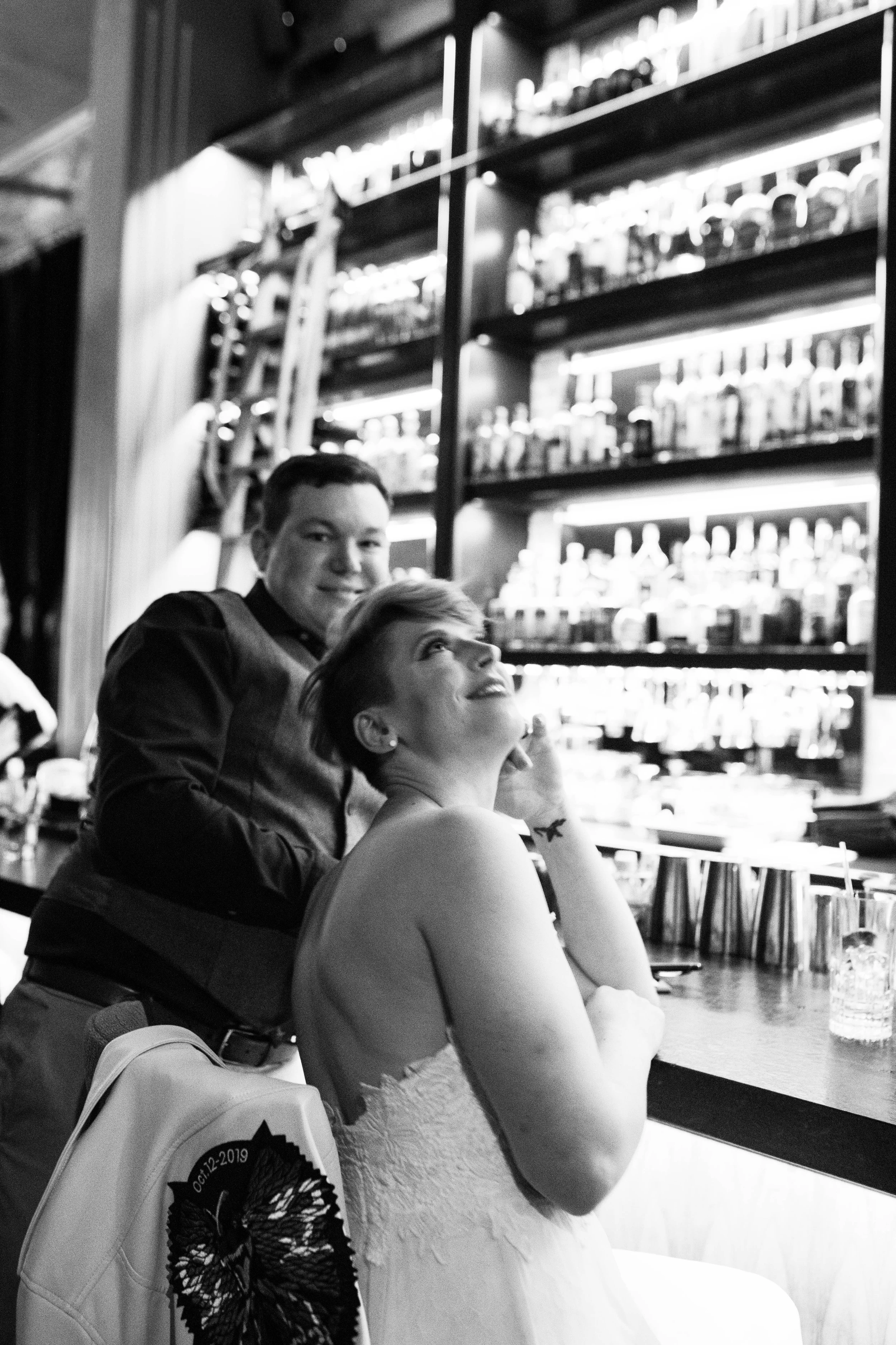 Black and white photo of a woman sitting at a bar with her elbow on the counter, looking up and smiling, with a man standing behind her smiling, in front of a backlit shelf of bottles. Pioneer Square, Seattle, WA wedding photography.