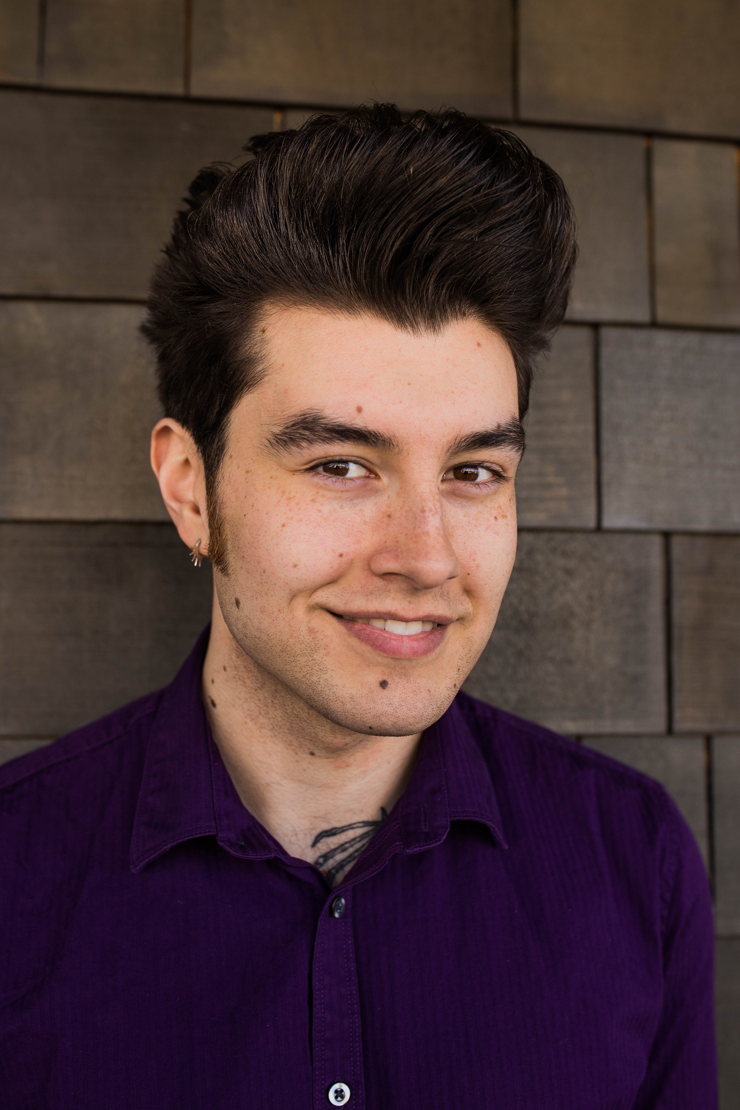 Close-up of a young man with dark hair in a pompadour style, wearing a purple shirt, smiling slightly, standing against a gray brick wall. Seattle professional head shot photography
