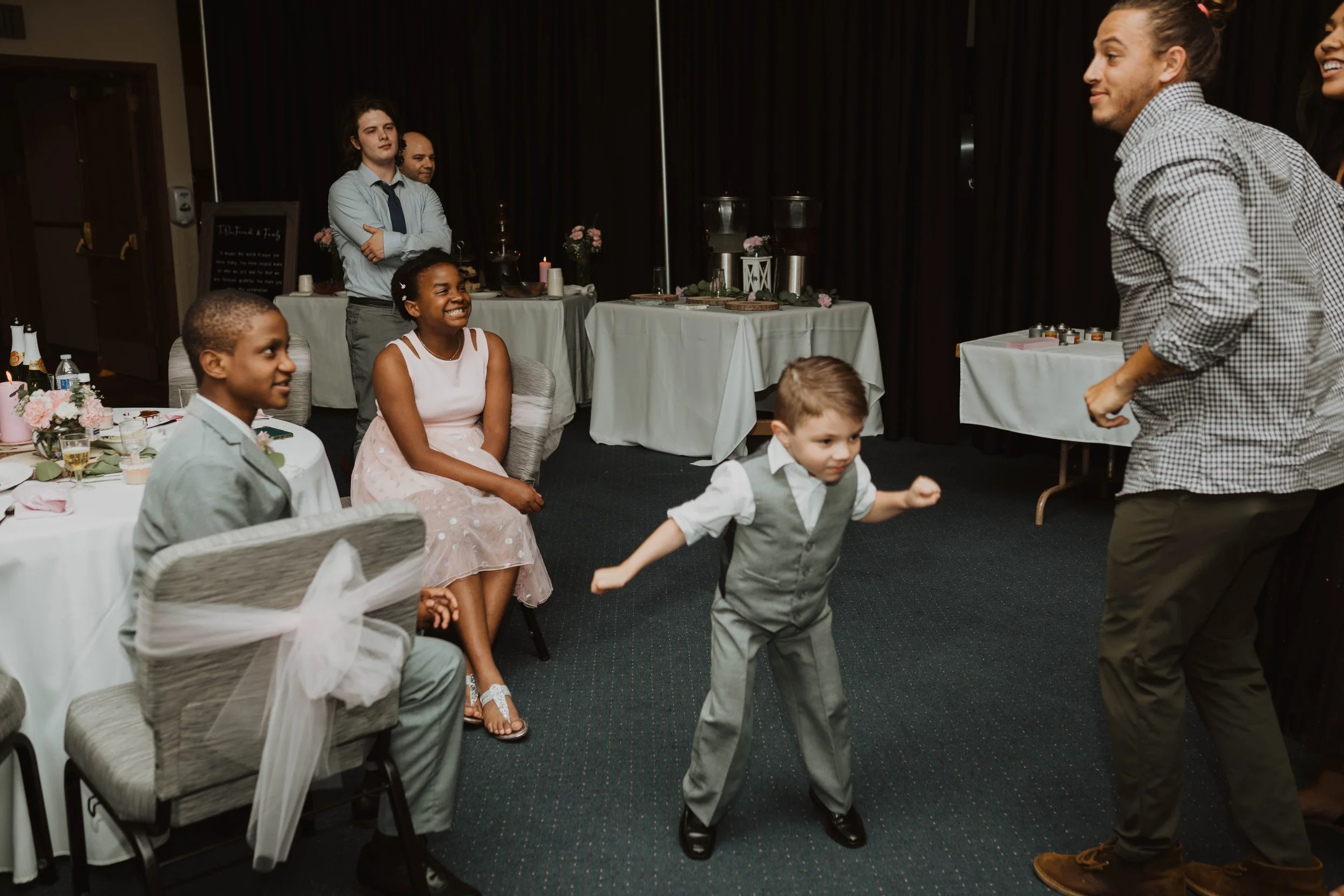 Young children dancing and smiling at a formal event, with adults sitting at decorated tables in the background. Seattle, WA wedding photography.