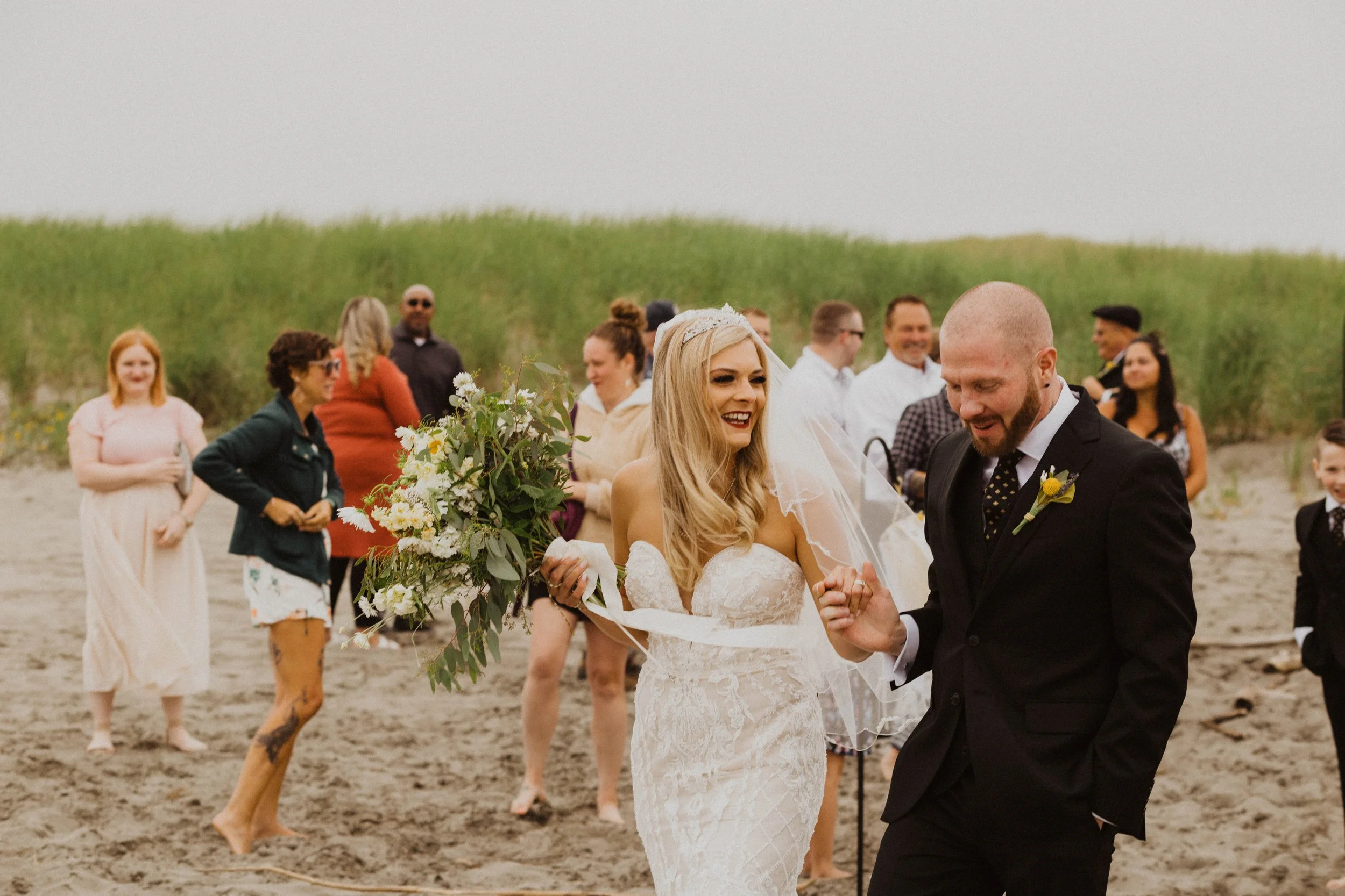 Bride and groom celebrating on the beach with friends and family, surrounded by sand dunes and cloudy sky. Long Beach, WA wedding photography.