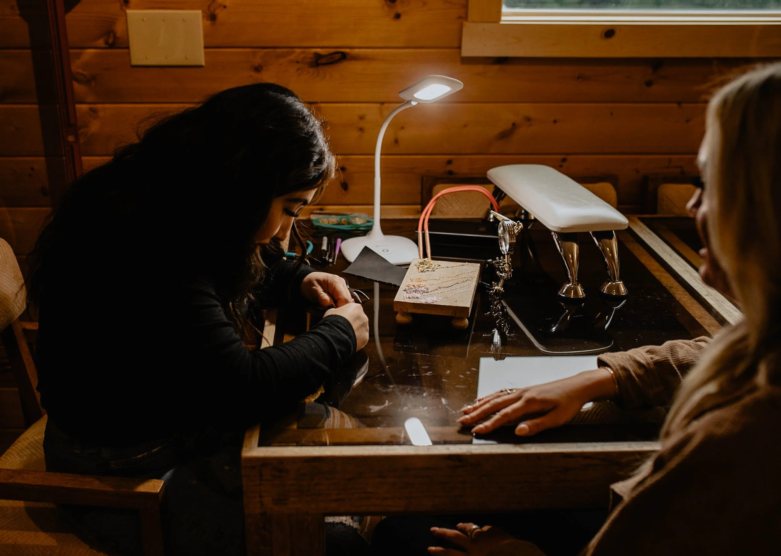 Two women sitting at a wooden table in a cozy, wood-paneled room, working on jewelry with various tools and jewelry displays on the table. Seattle professional head shot photography