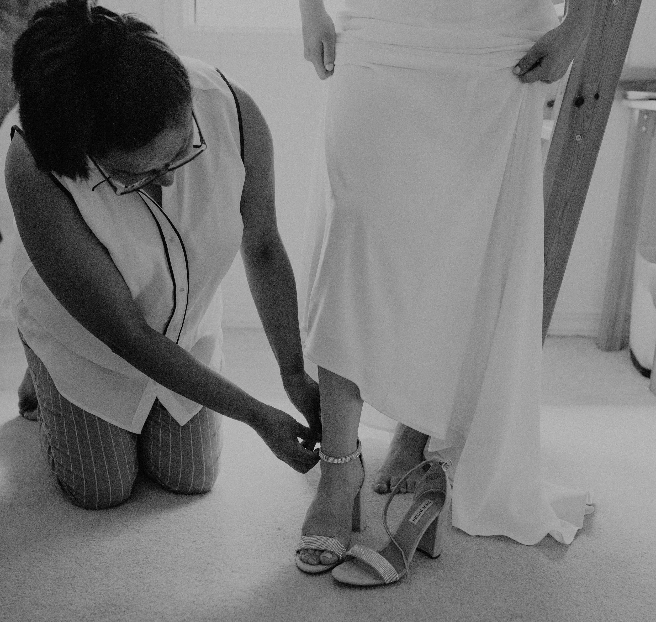 A woman kneeling down and fastening the ankle strap of a high-heeled shoe on another person in a white dress. Seattle, WA wedding photography.