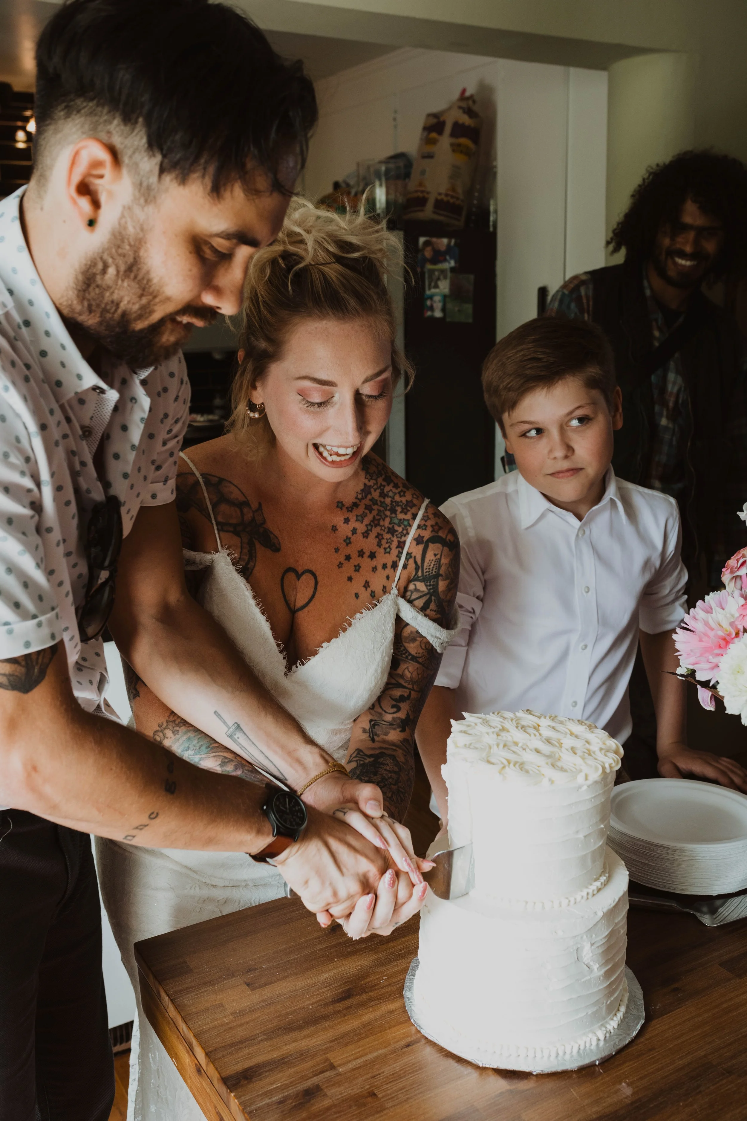 A couple with tattoos and a wedding dress cut a wedding cake together, smiling, while a child in a white shirt watches. Seattle, WA wedding photography.