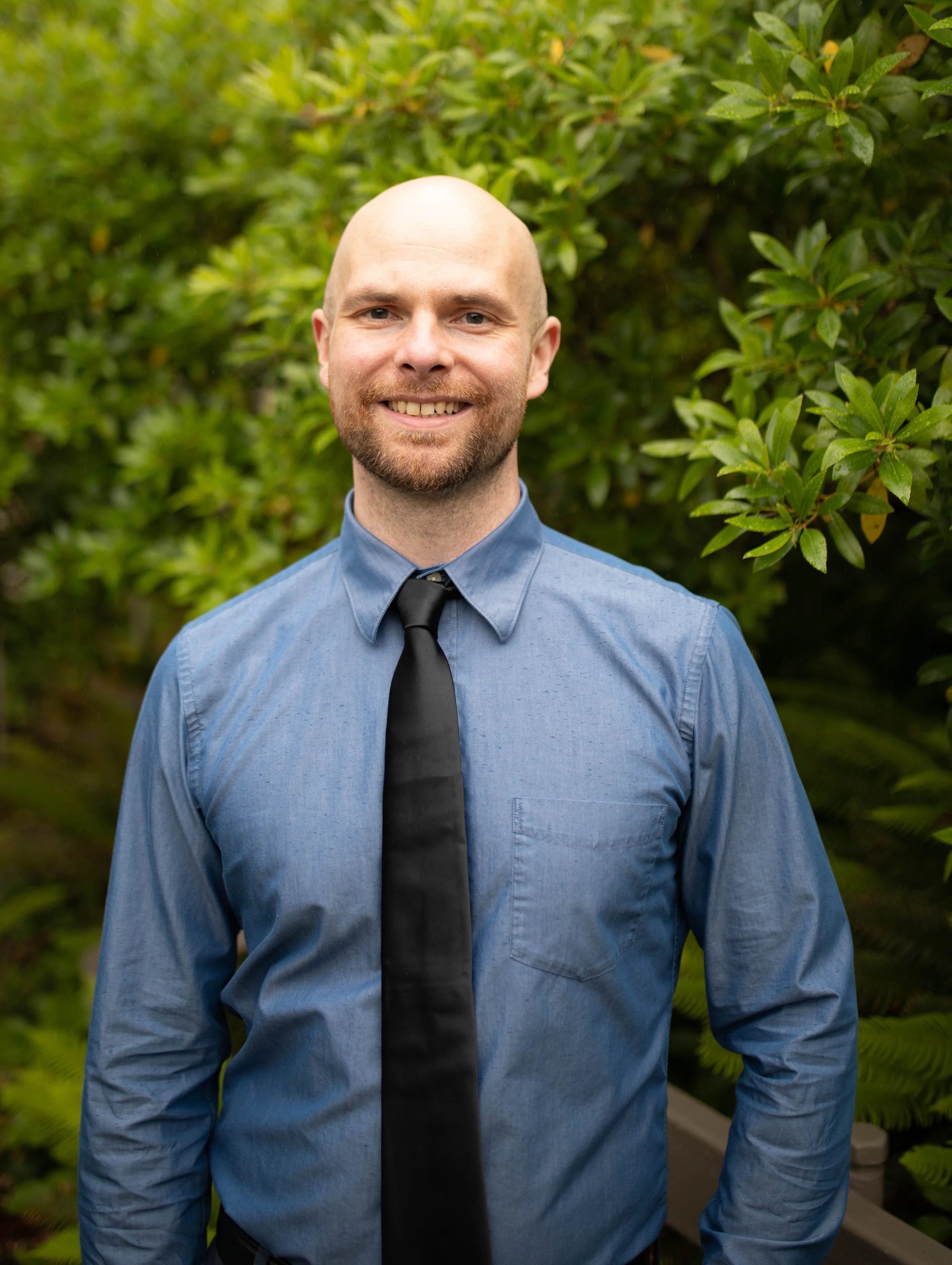 Smiling man with a beard and shaved head standing outdoors in front of green foliage, wearing a blue button-up shirt and a black tie. Seattle professional head shot photography
