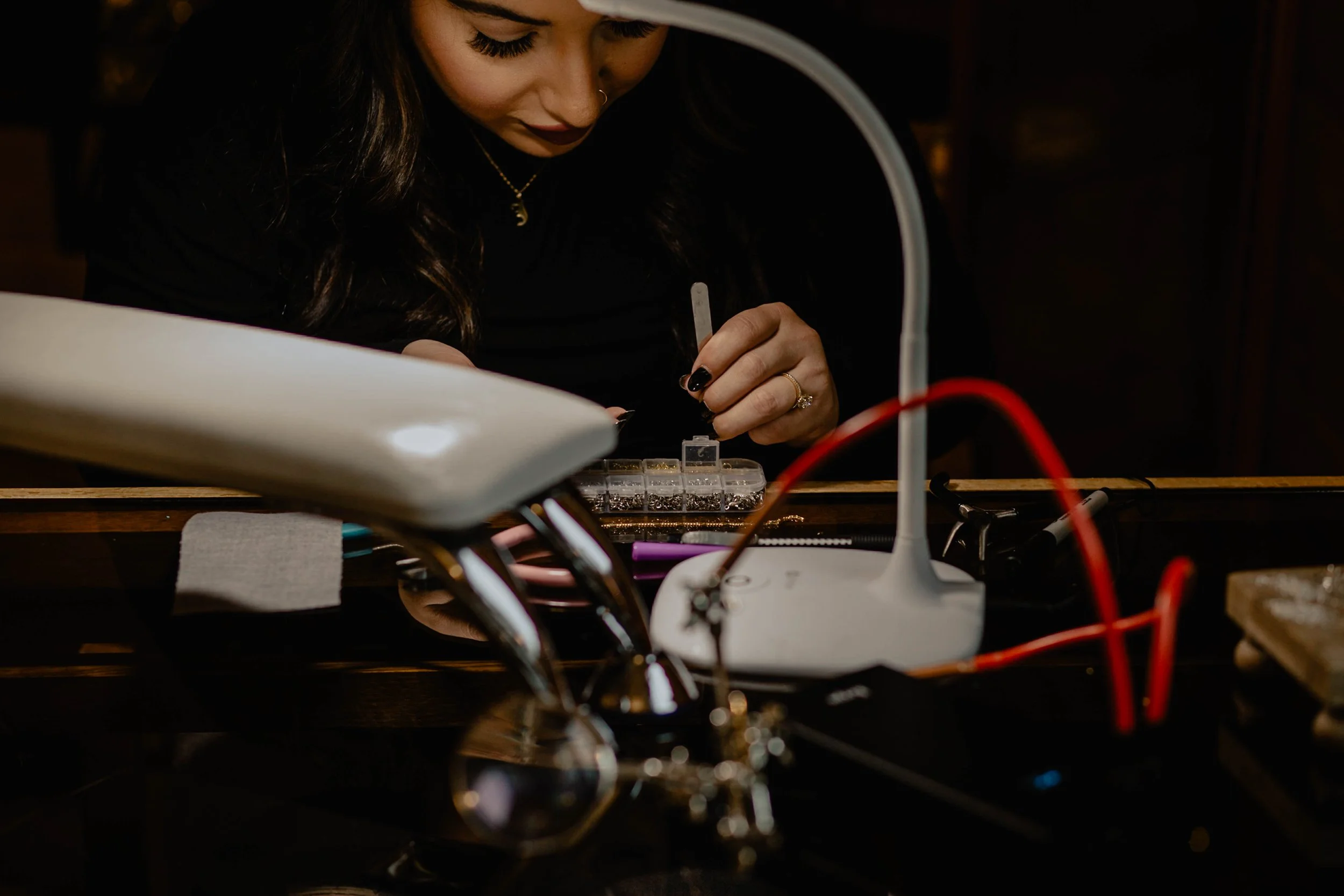A woman with dark hair and dark lipstick working with small tools and beads at a table, surrounded by electronic devices and jewelry-making supplies. Seattle professional head shot photography