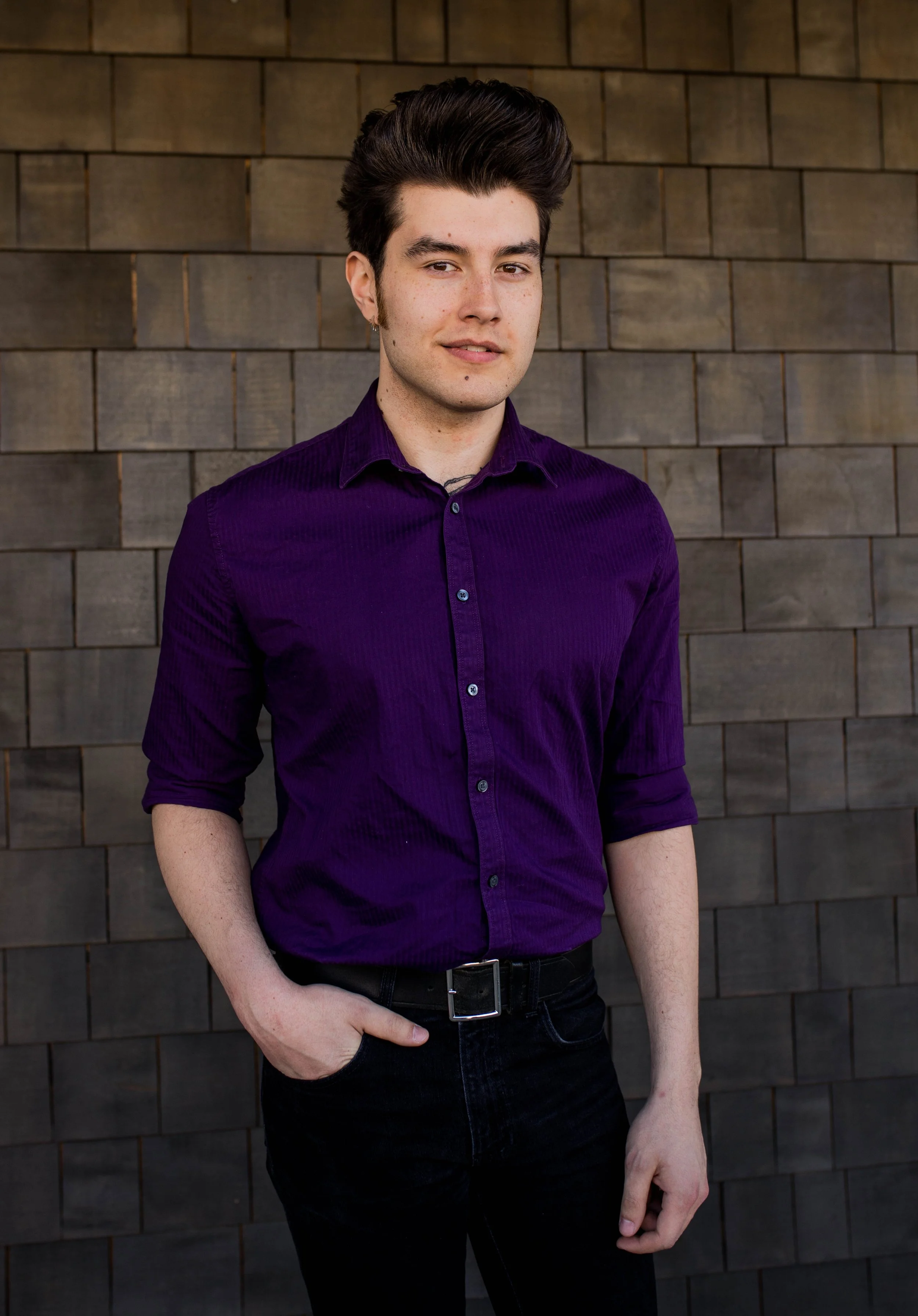 A young man with dark, styled hair, wearing a purple button-up shirt and black pants, standing against a dark wooden wall. Seattle professional head shot photography