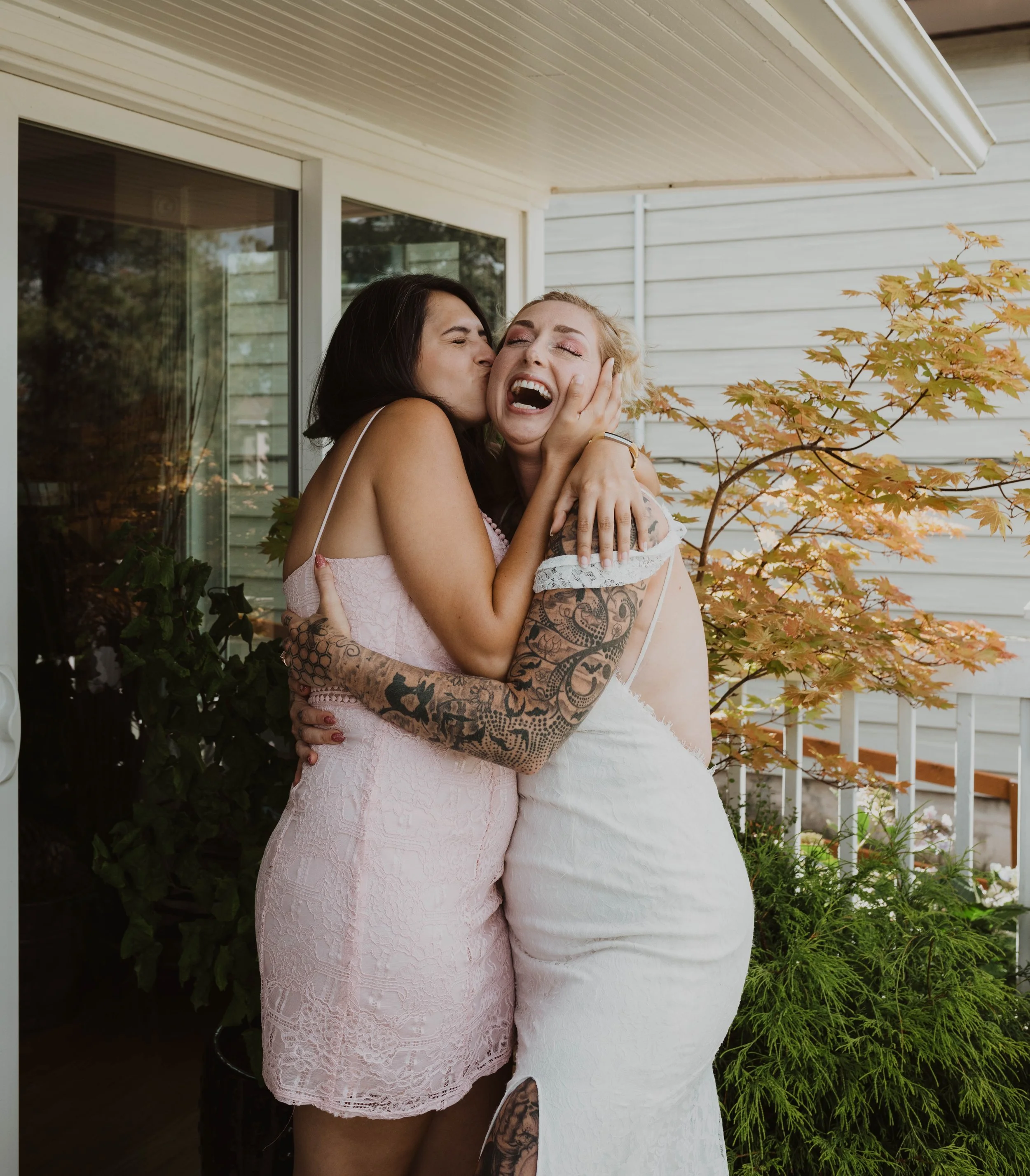 Two women embracing and laughing on a porch, with one kissing the other's cheek, surrounded by trees and plants. Seattle, WA wedding photography.
