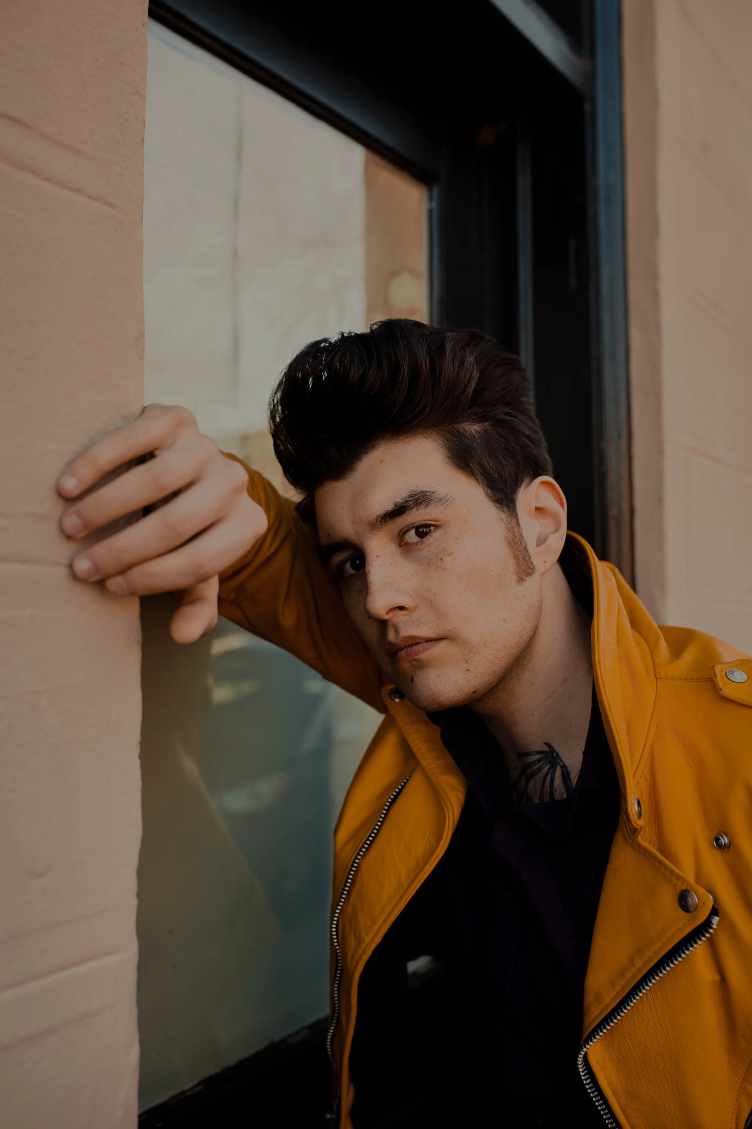 Young man with dark hair, freckles, and a tattoo on his neck, wearing a yellow jacket, leaning against a wall with one arm raised, near a window. Seattle professional head shot photography