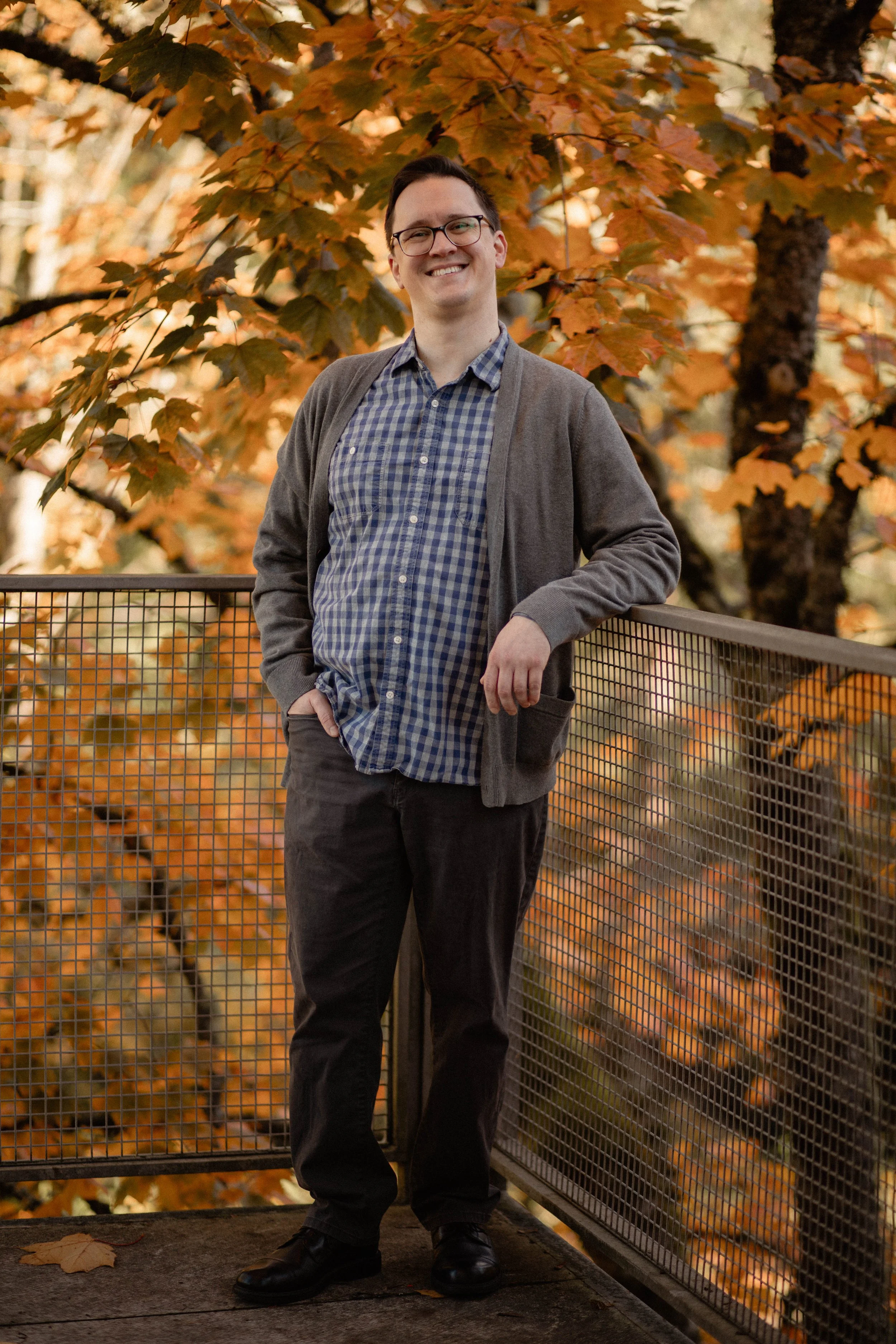 A smiling man wearing glasses, a checkered shirt, and a gray cardigan standing on a balcony with autumn leaves in the background. Seattle professional head shot photography
