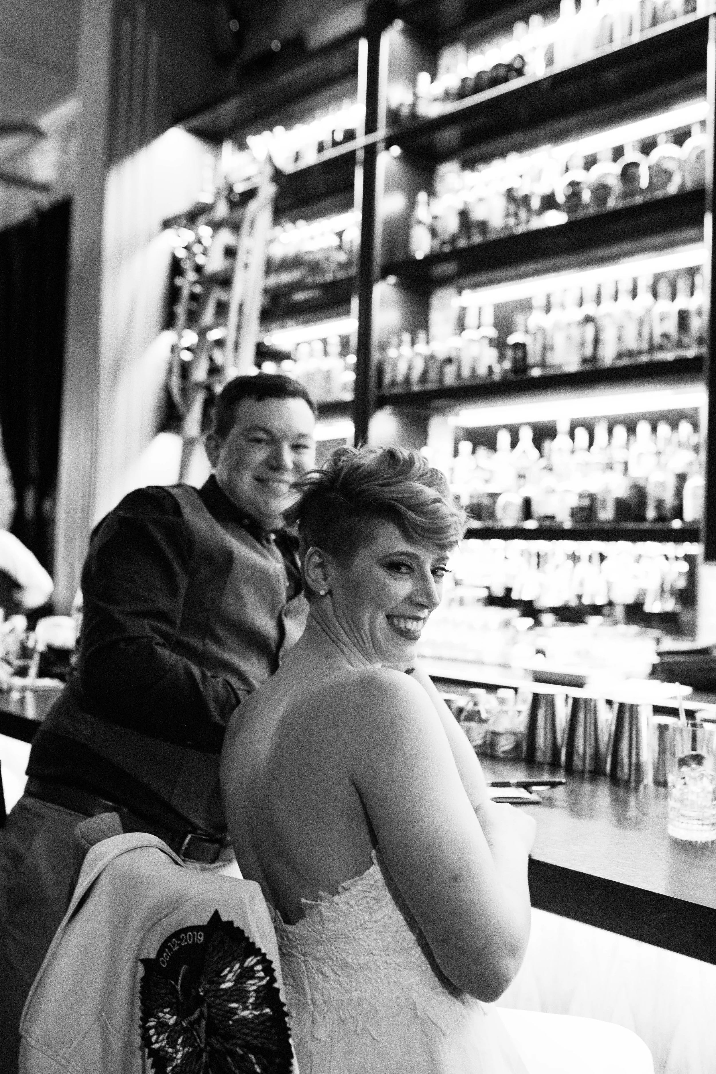 A black and white photo of a smiling woman in a strapless dress sitting at a bar counter, with a man standing behind her. The background shows a shelf filled with bottles and a ladder leaning against the wall.