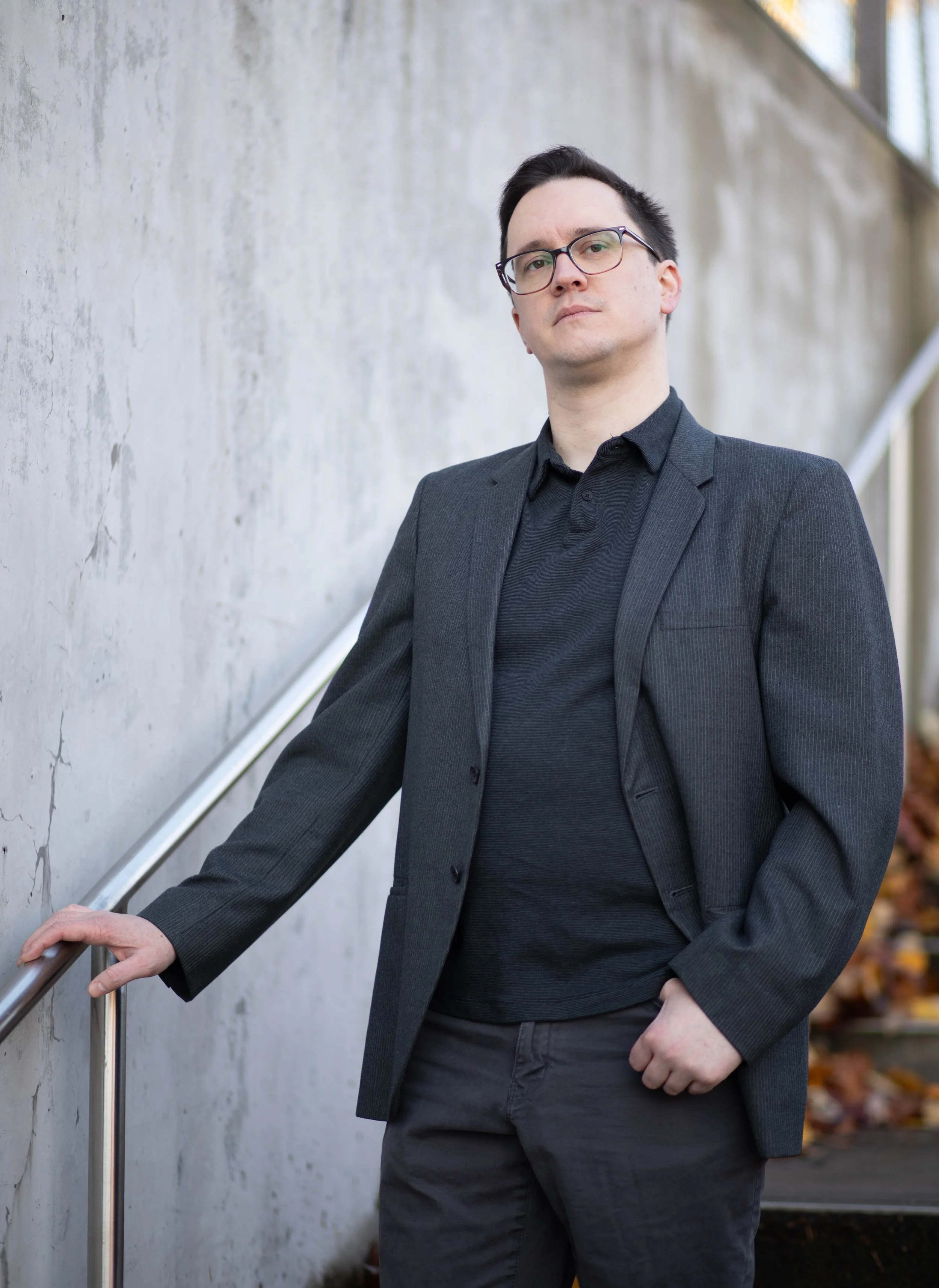 A young man in a dark blazer and glasses standing outdoors beside a concrete wall. Seattle professional head shot photography