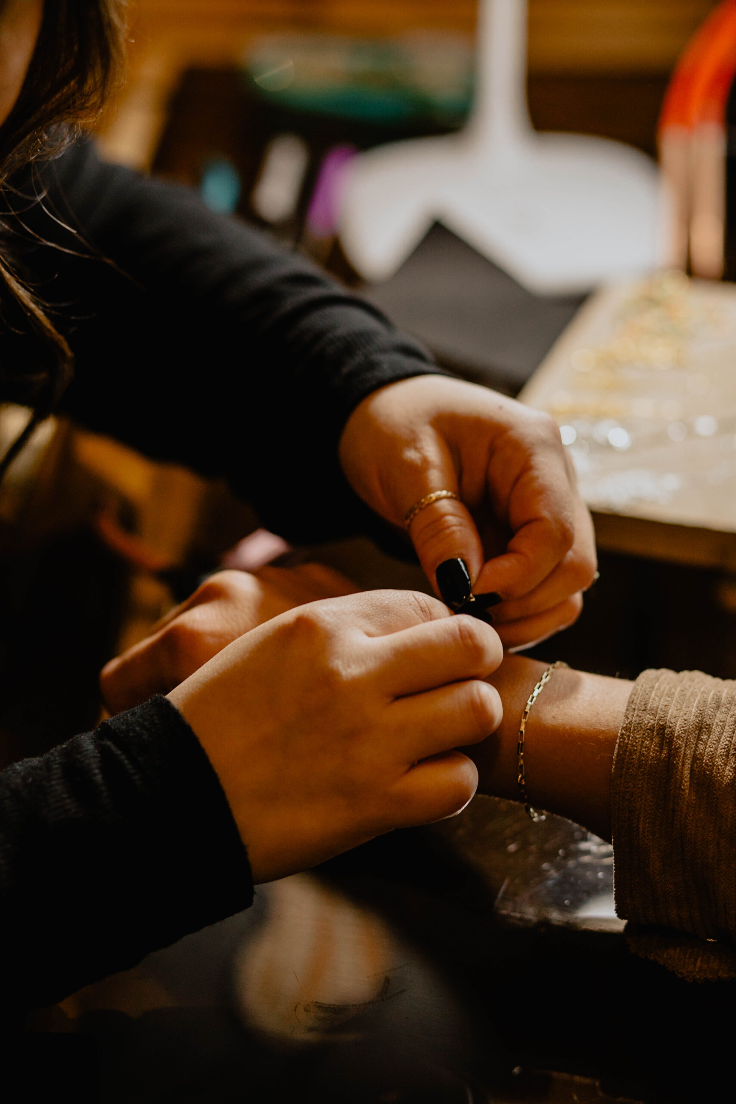 Close-up of a person jewelry-making, focusing on hands. One person is holding a bracelet, while the other person is assisting or adjusting it. The background is slightly blurred with a decorative star and craft supplies visible. Seattle professional 