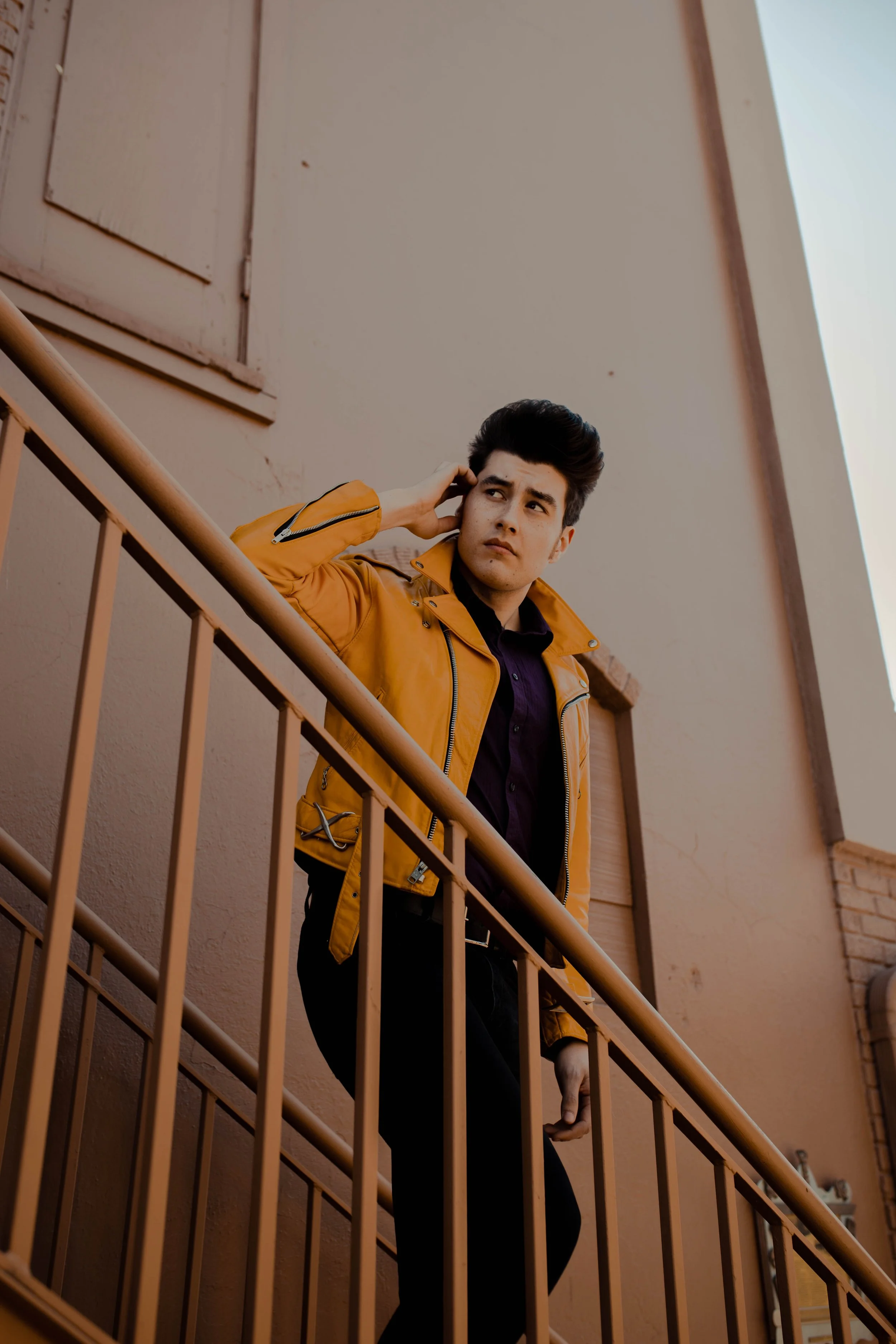 Young man with dark hair, wearing a yellow leather jacket and black pants, standing on a balcony, looking thoughtfully into the distance with his hand near his ear. Seattle professional head shot photography
