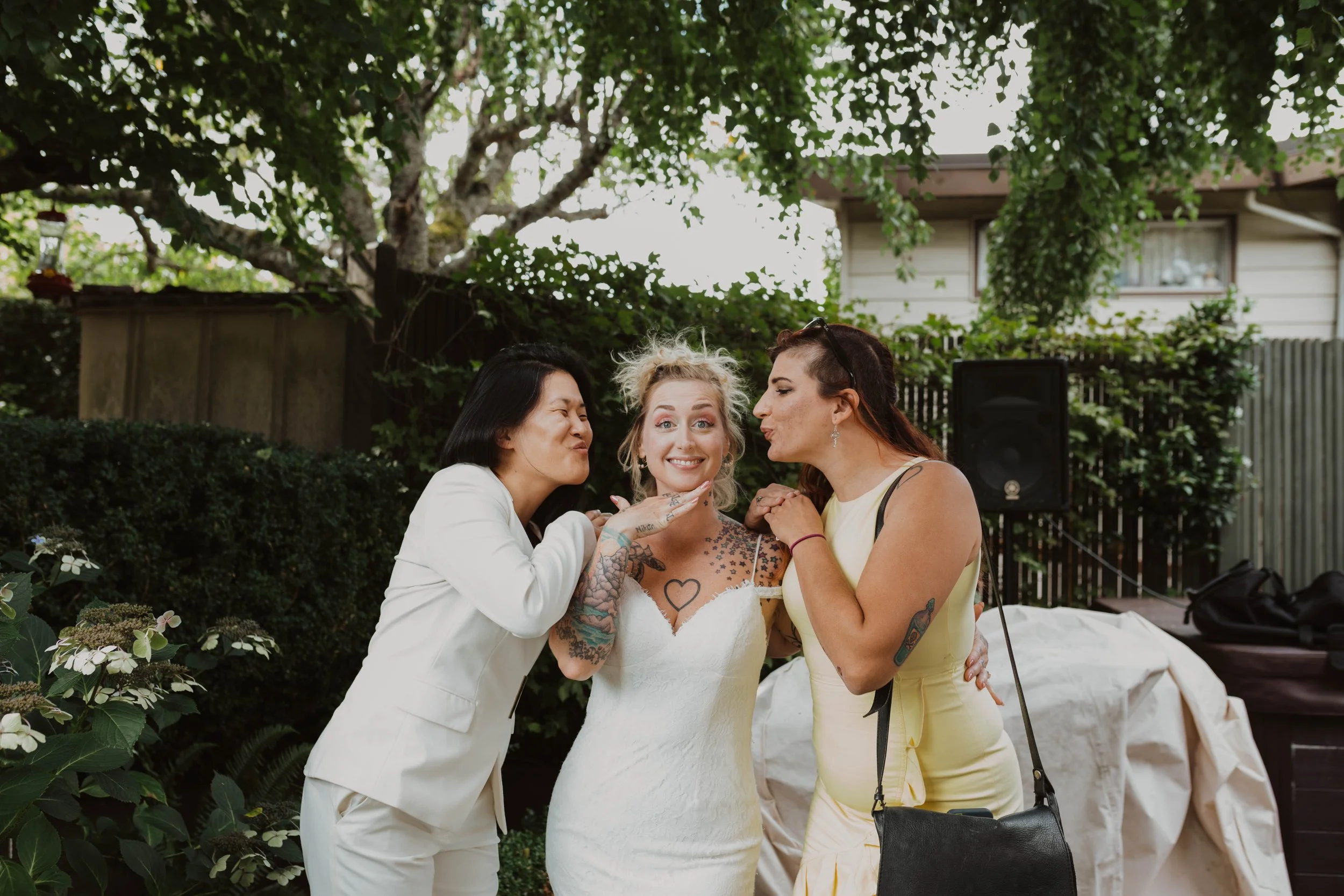 Three women, one in a white dress with tattoos and two others in light-colored outfits, are outside under trees. They are smiling and making playful faces directed at each other. Seattle, WA wedding photography.