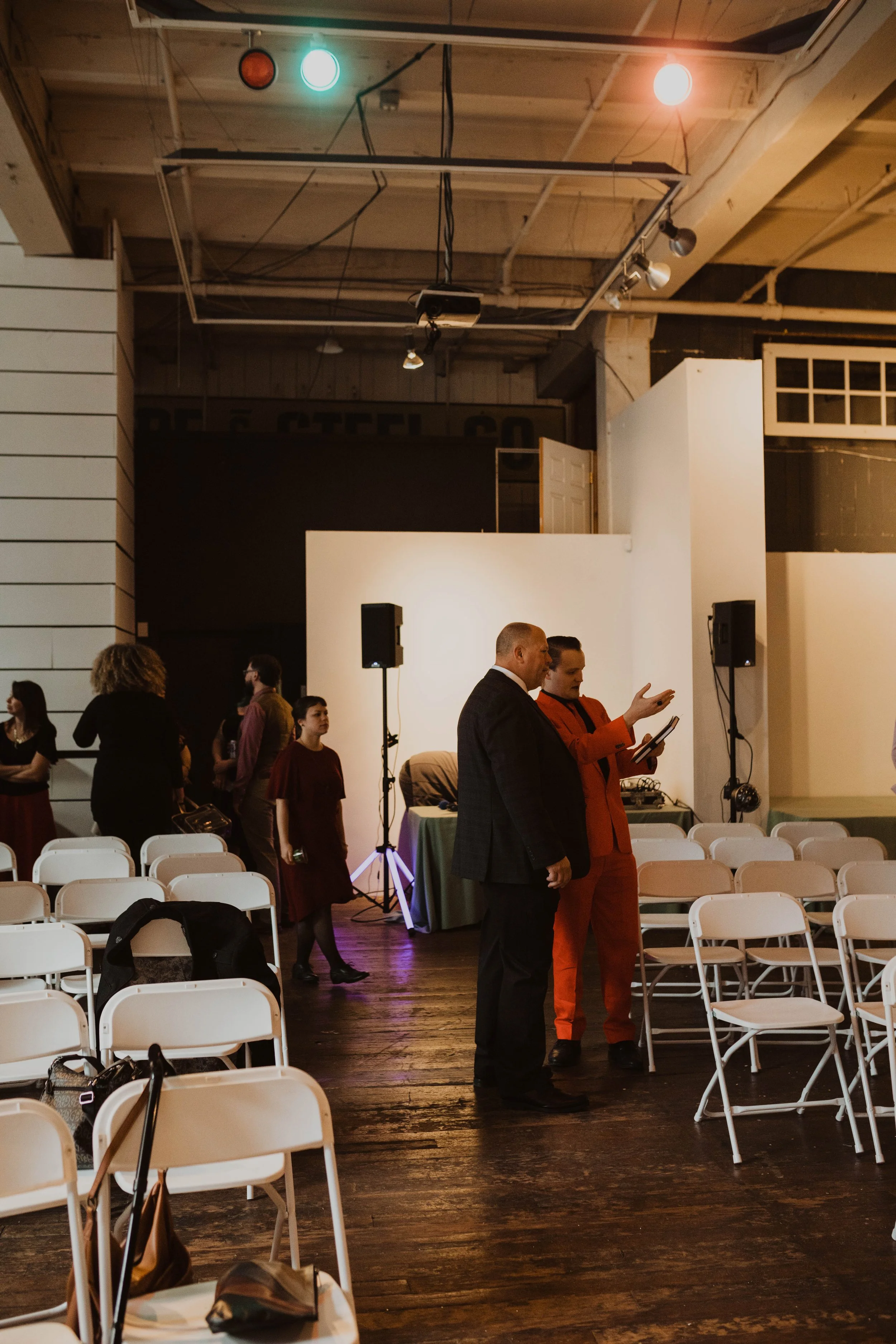 People gathering at an indoor event space with chairs, some are engaging in conversations and preparing for a presentation or performance. The room has a rustic wooden floor, white walls, and stage lighting. Pioneer Square, Seattle, WA wedding photog