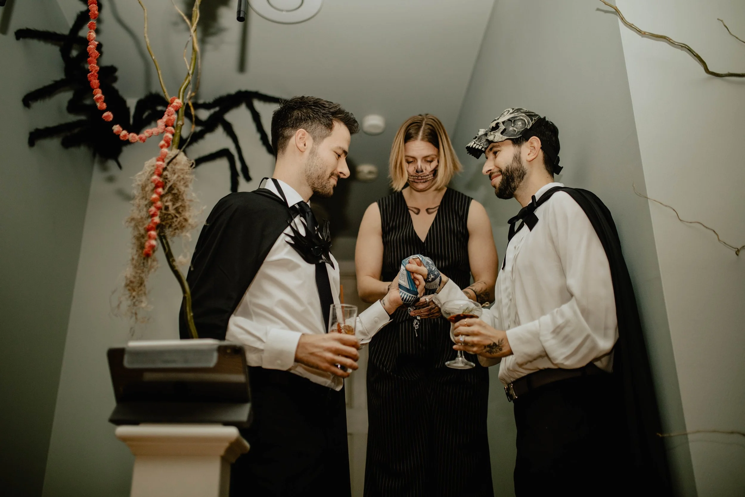 Two men dressed as Dracula and Frankenstein shake hands while a woman in a skeleton costume looks on at a Halloween party decorated with hanging spiders and spider webs. Seattle wedding photography.