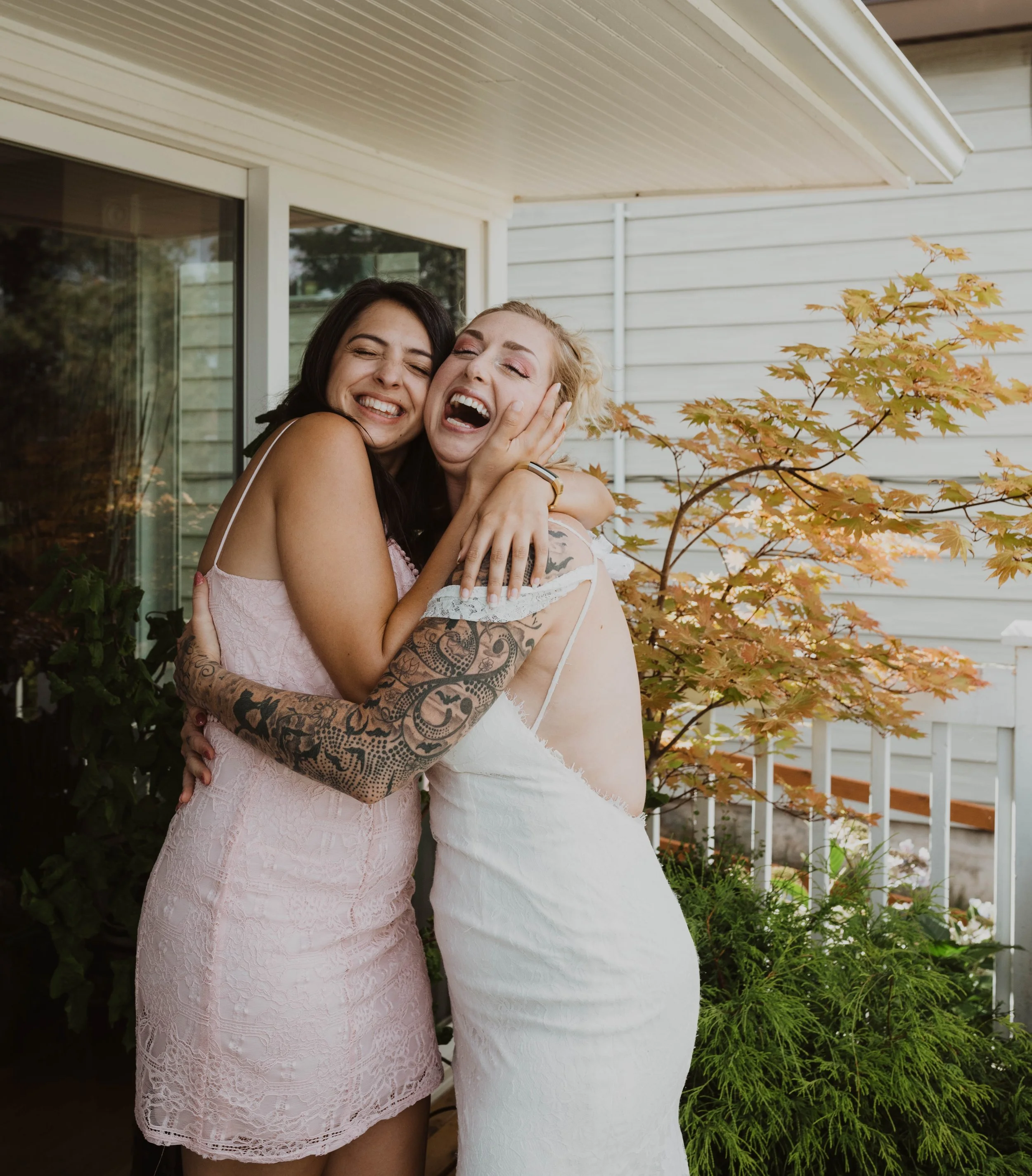 Two women embracing and laughing on a porch, one in a pink dress, the other in a white dress, with a Japanese maple tree and white house siding in the background. Seattle, WA wedding photography.