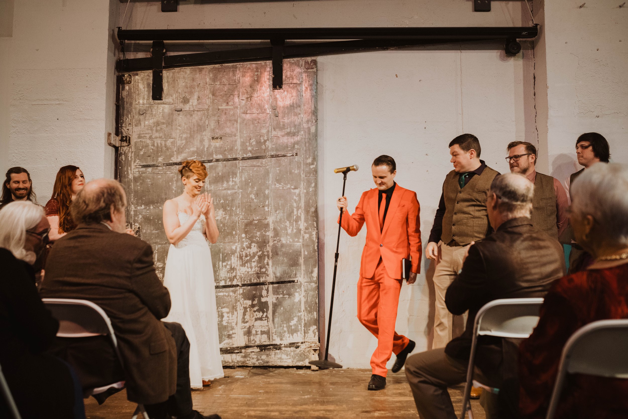 A wedding ceremony with the bride in a white dress and the groom in a red suit standing in front of an audience in an industrial-style venue. The groom is holding a microphone and looking down, while the bride has her hands together and appears happy