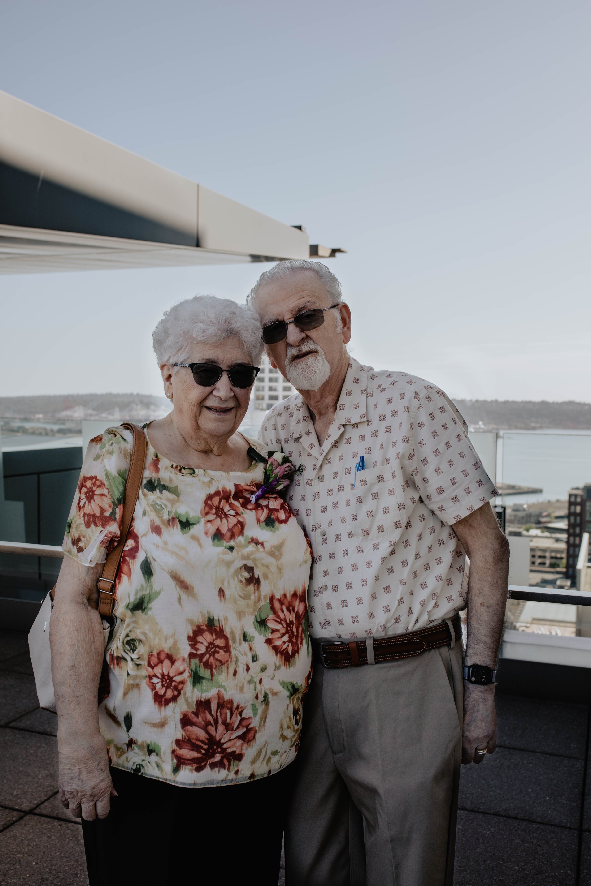 An elderly couple wearing sunglasses and smiling on a balcony with a city skyline and water in the background. Seattle Municipal Courthouse wedding photography.