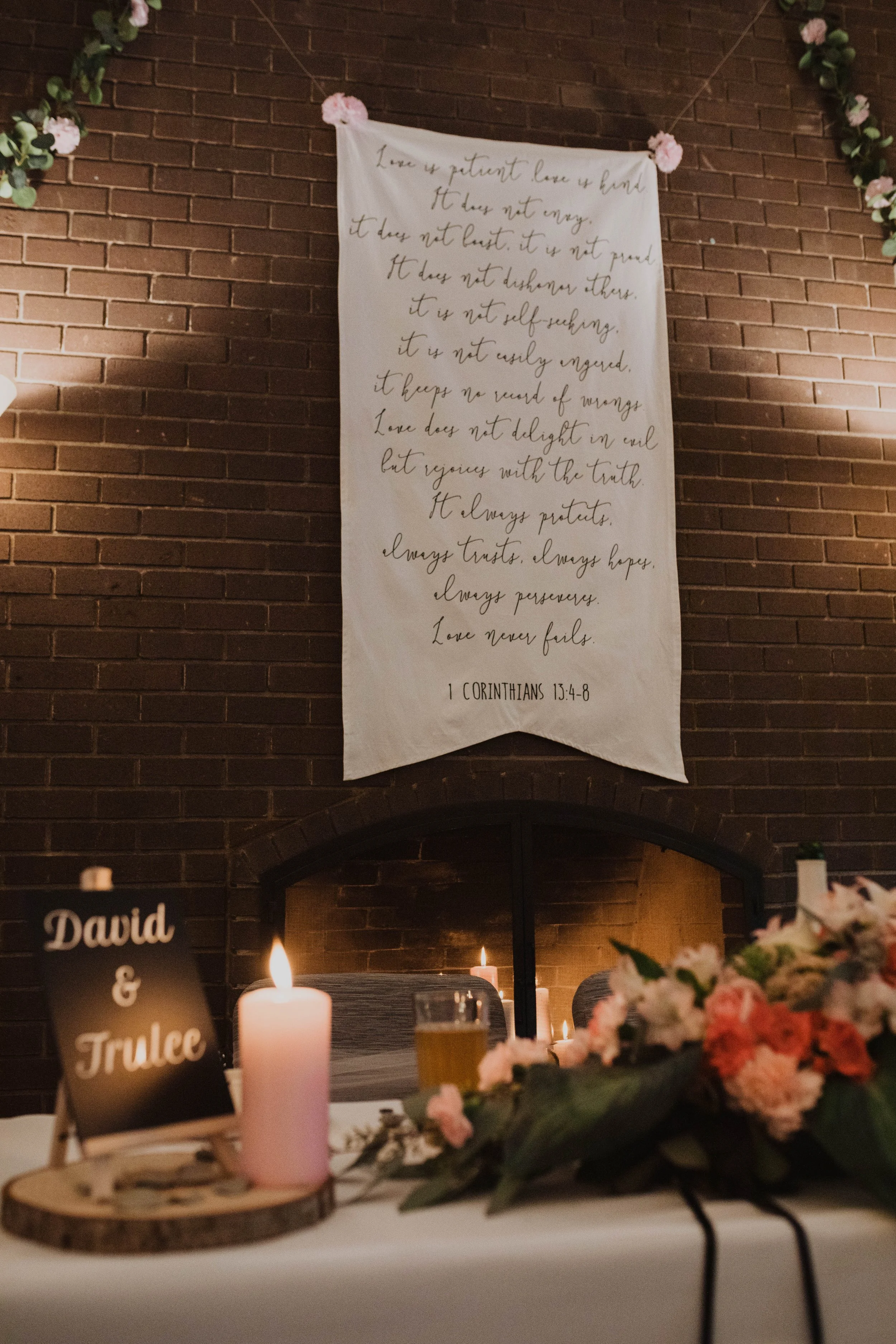Wedding reception with a table decorated with flowers, a candle, and a sign that reads 'David & Trulee'. In the background, a large banner displays a Christian scripture from 1 Corinthians 13:4-8, hanging on a brick wall with pink floral decorations.