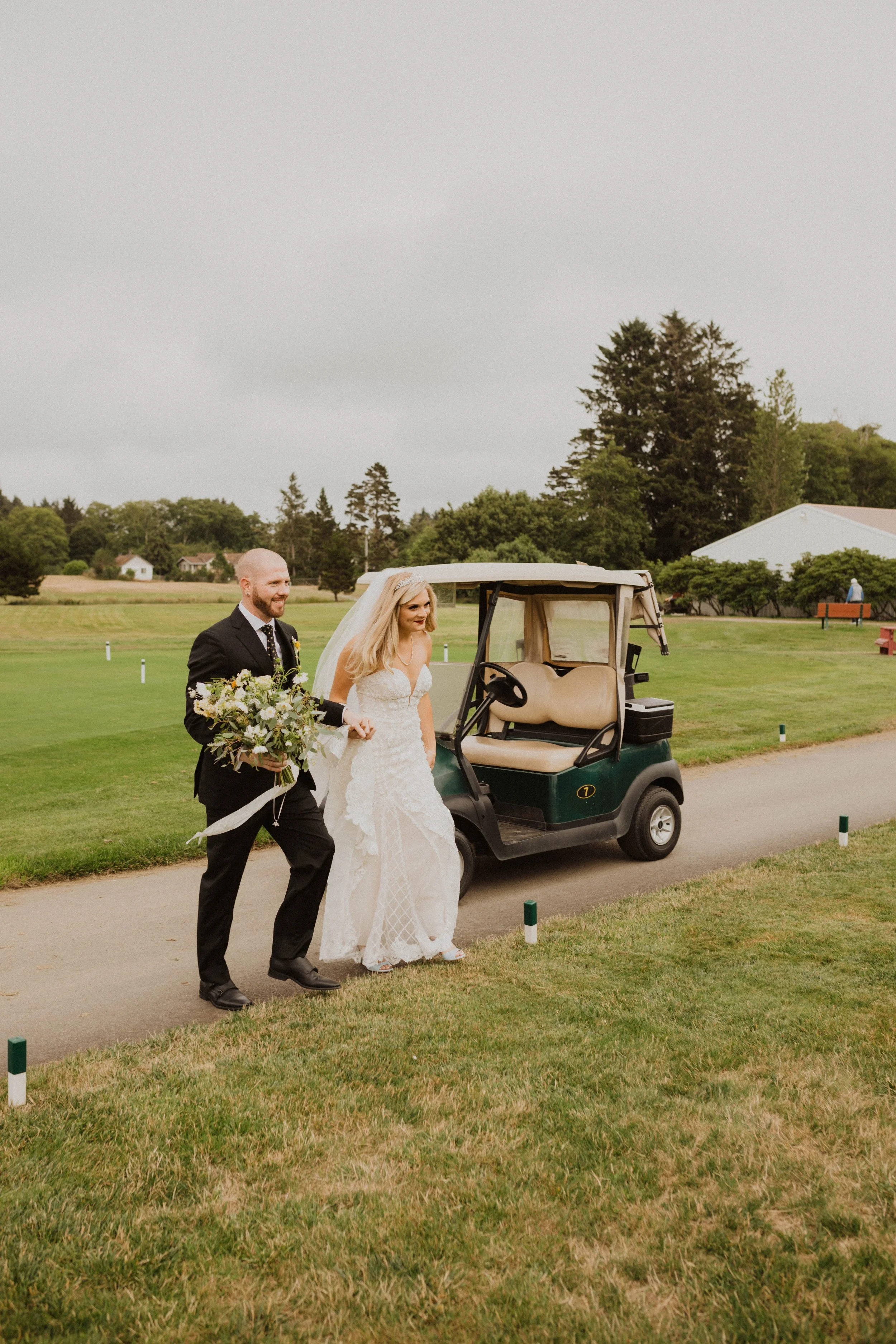 A bride and groom walking hand in hand on a golf course, with the bride holding a bouquet of flowers and the groom dressed in a black suit, next to a golf cart. The sky is overcast. Long Beach, WA wedding photography.
