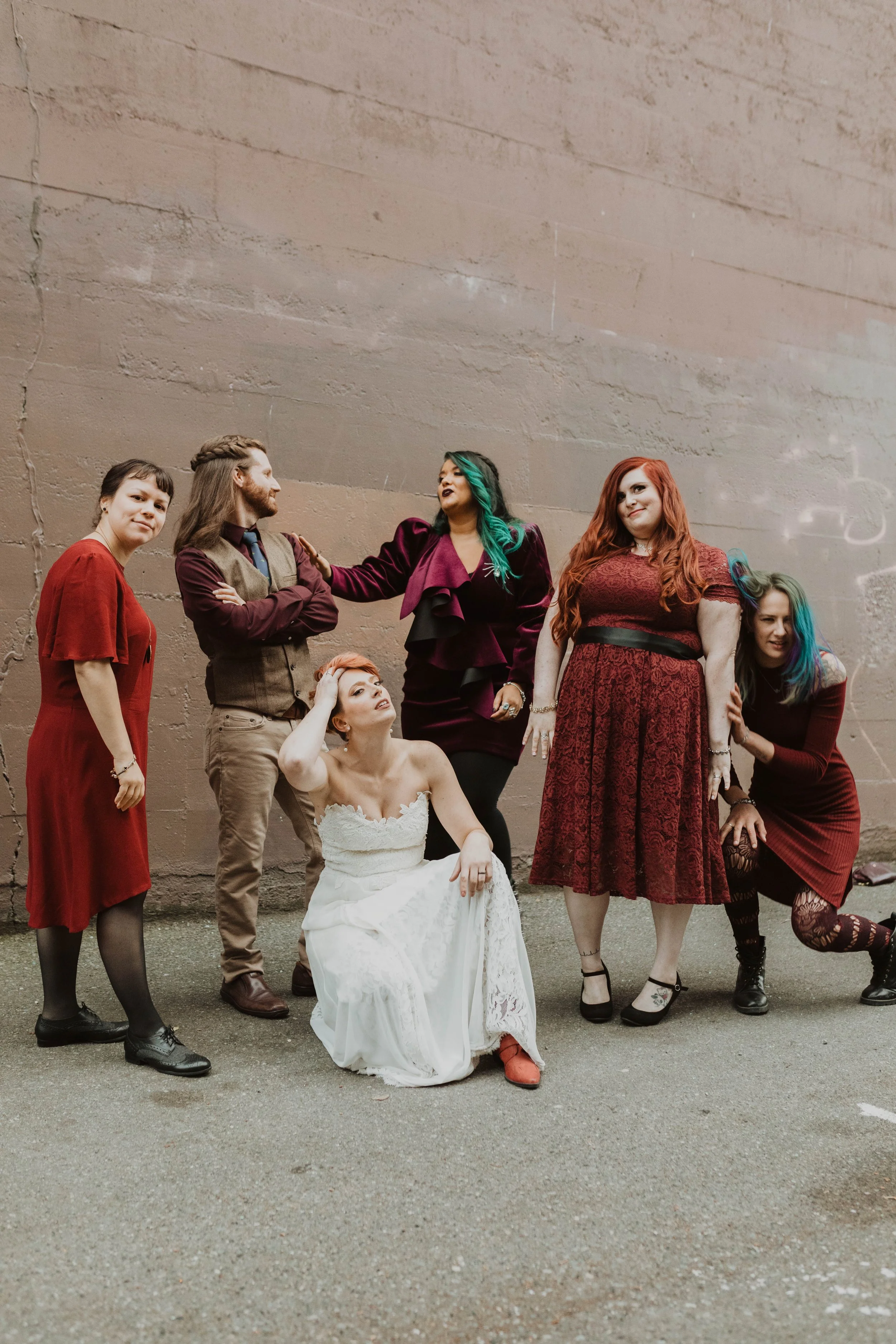 Group of six women with colorful hair and diverse outfits posing against a textured wall, with one woman in a white dress sitting on the ground. Pioneer Square, Seattle, WA wedding photography.