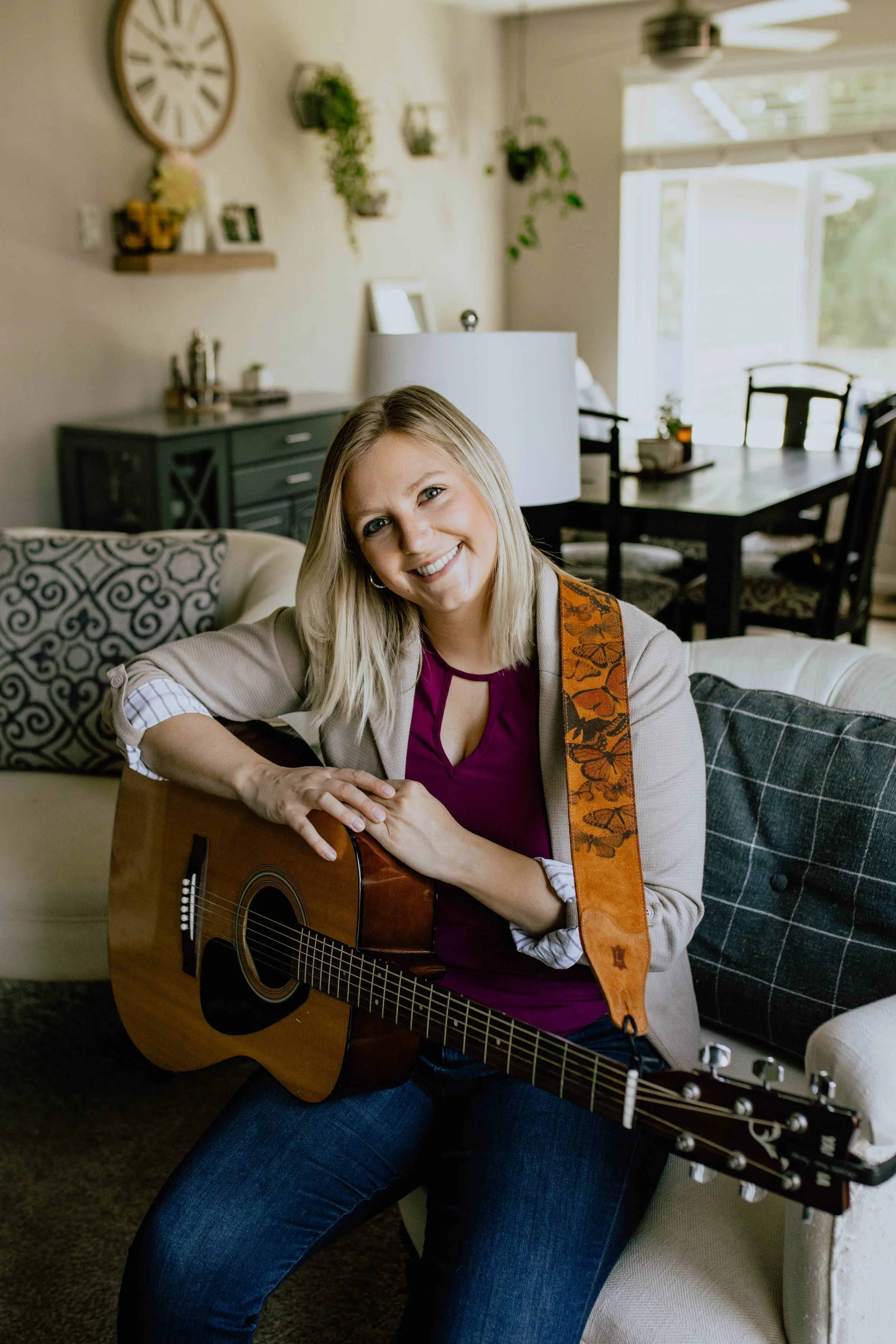 A woman sitting on a beige sofa holding an acoustic guitar, smiling in a living room with plants and a dining table in the background. Seattle professional head shot photography