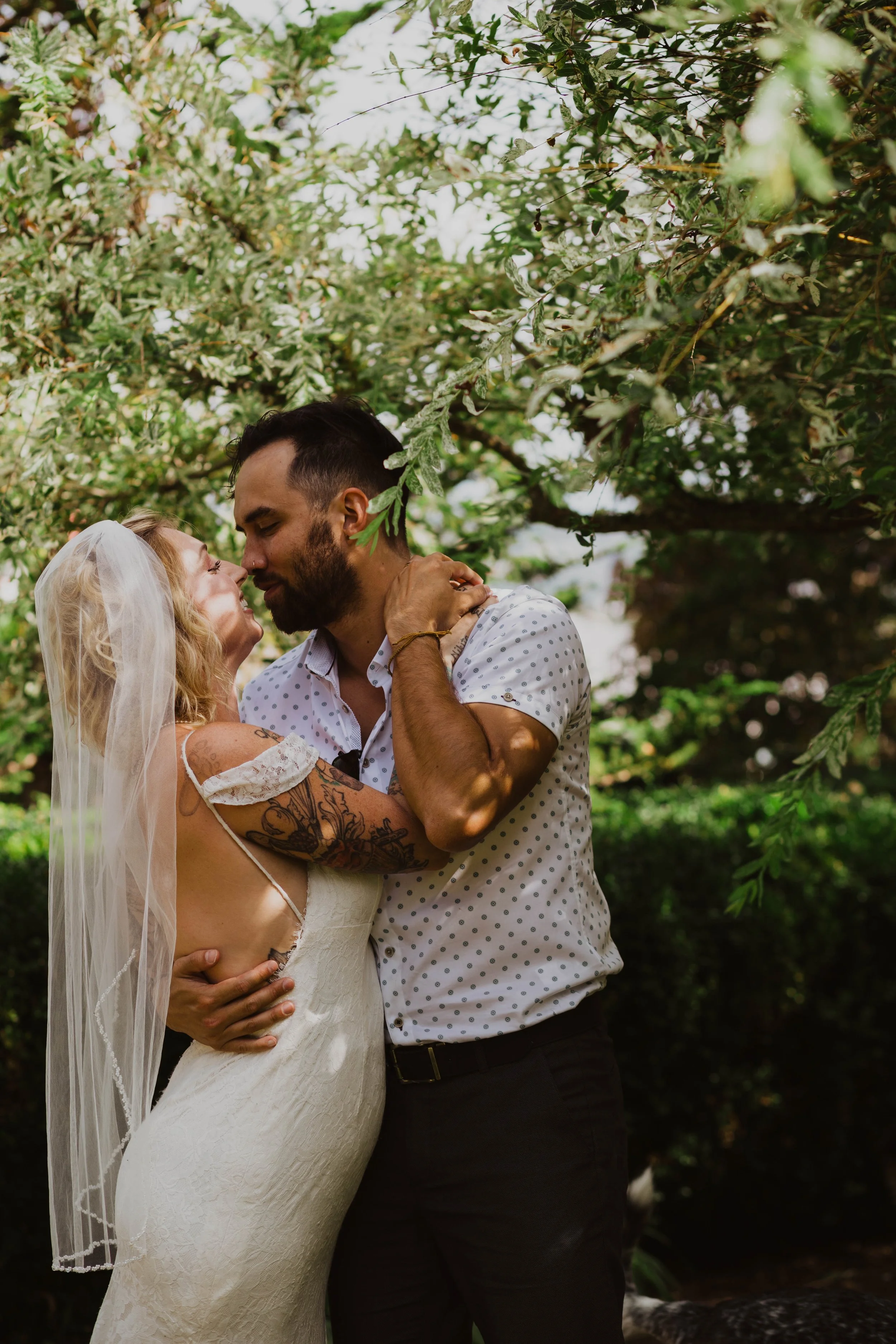 A couple embracing outdoors under a tree, with the woman wearing a wedding dress with a veil and the man in a short sleeve, polka dot shirt, both smiling  Seattle, WA wedding photography.