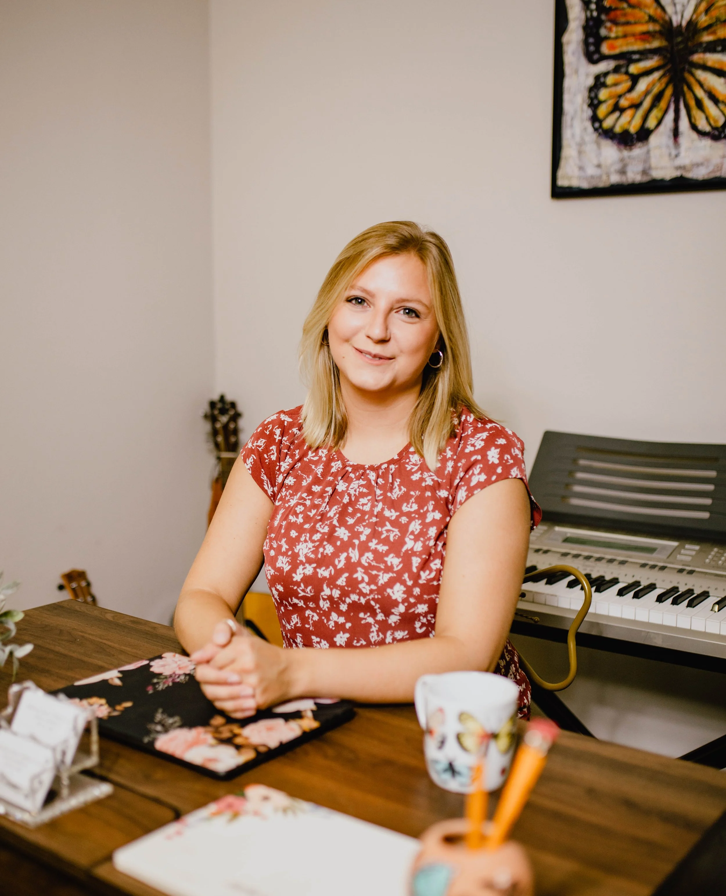 A woman with blonde hair in a red floral dress sitting at a wooden table with a notebook, mug, and other items, smiling at the camera. A digital keyboard is in the background, along with a colorful butterfly artwork on the wall. Seattle professional 