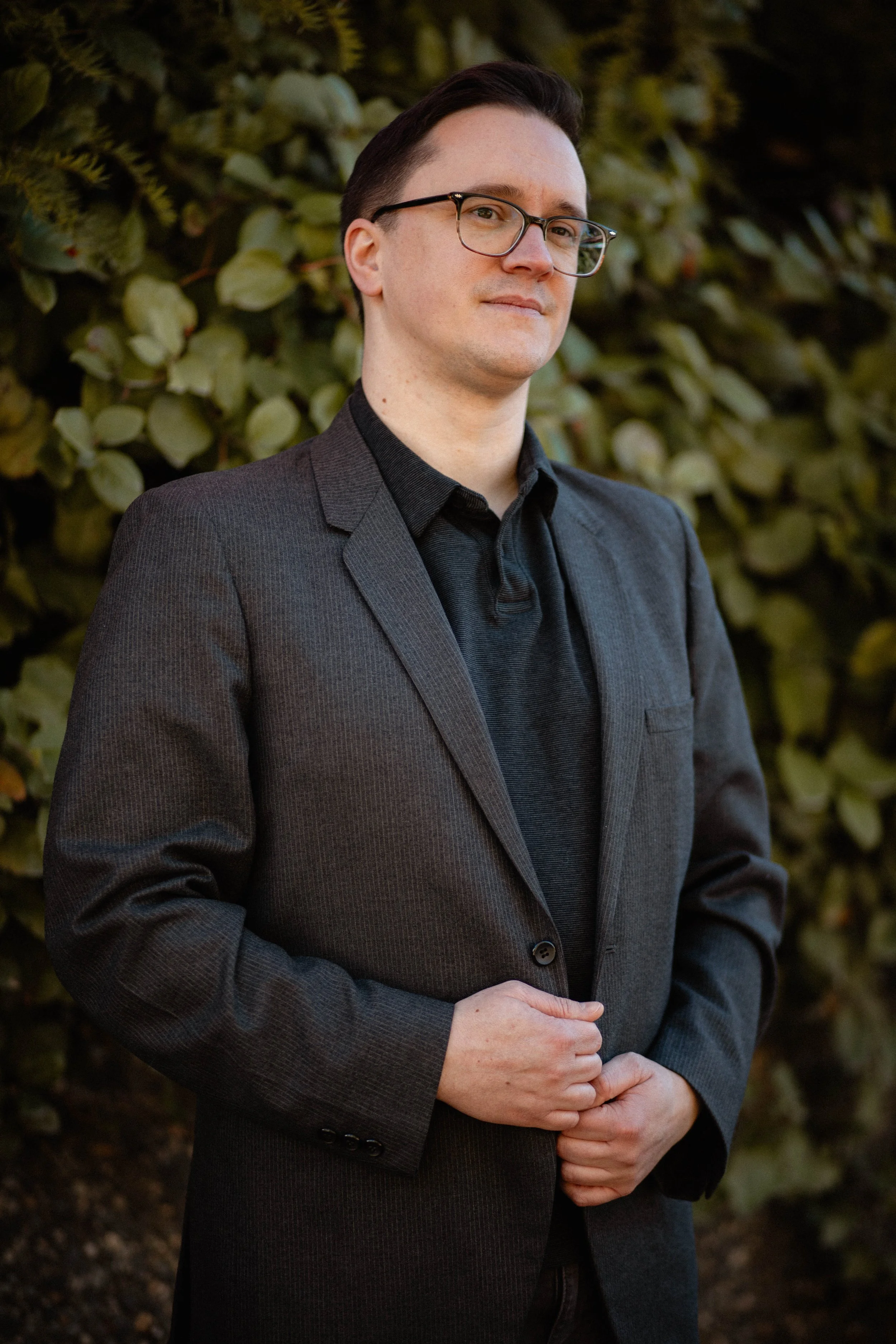 A man with glasses and dark hair, wearing a dark gray blazer and black shirt, standing outdoors in front of a leafy background. Seattle professional head shot photography