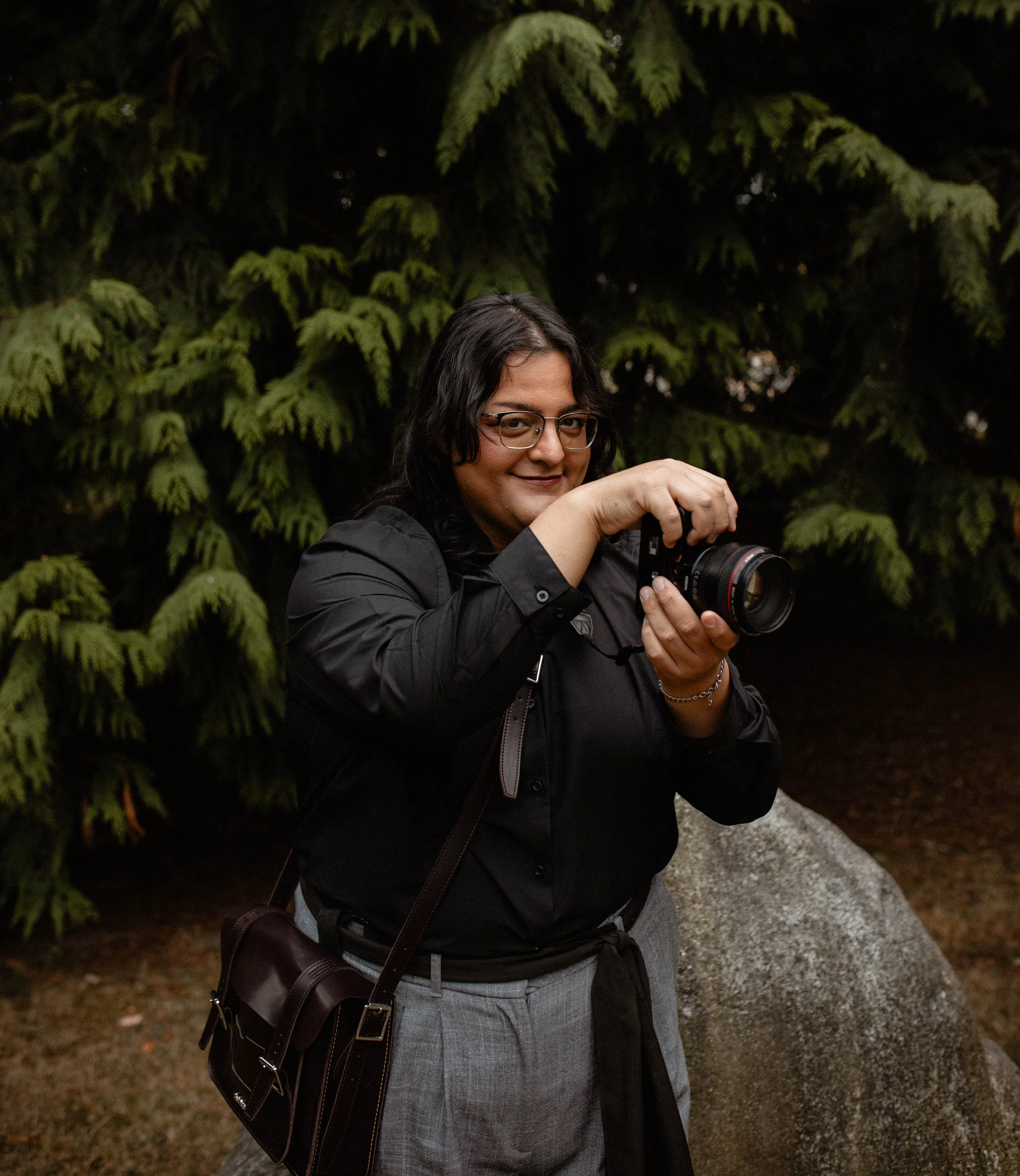 A woman with glasses holding a camera, standing outdoors near a large rock and green trees. Seattle professional head shot photography