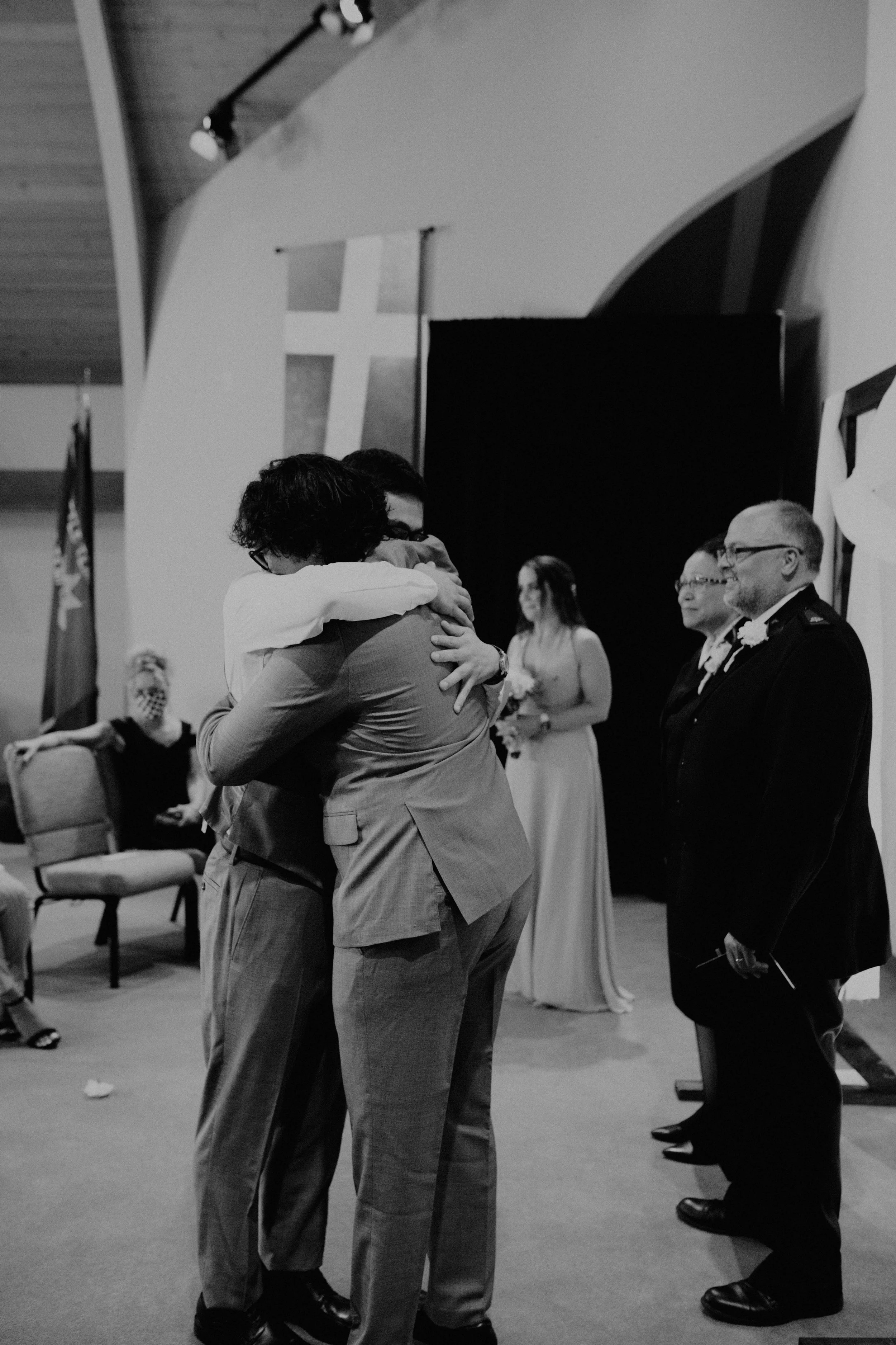 Two men embrace in a hug during a wedding ceremony, with the bride and officiant standing in the background, and guests seated nearby. Seattle, WA wedding photography.