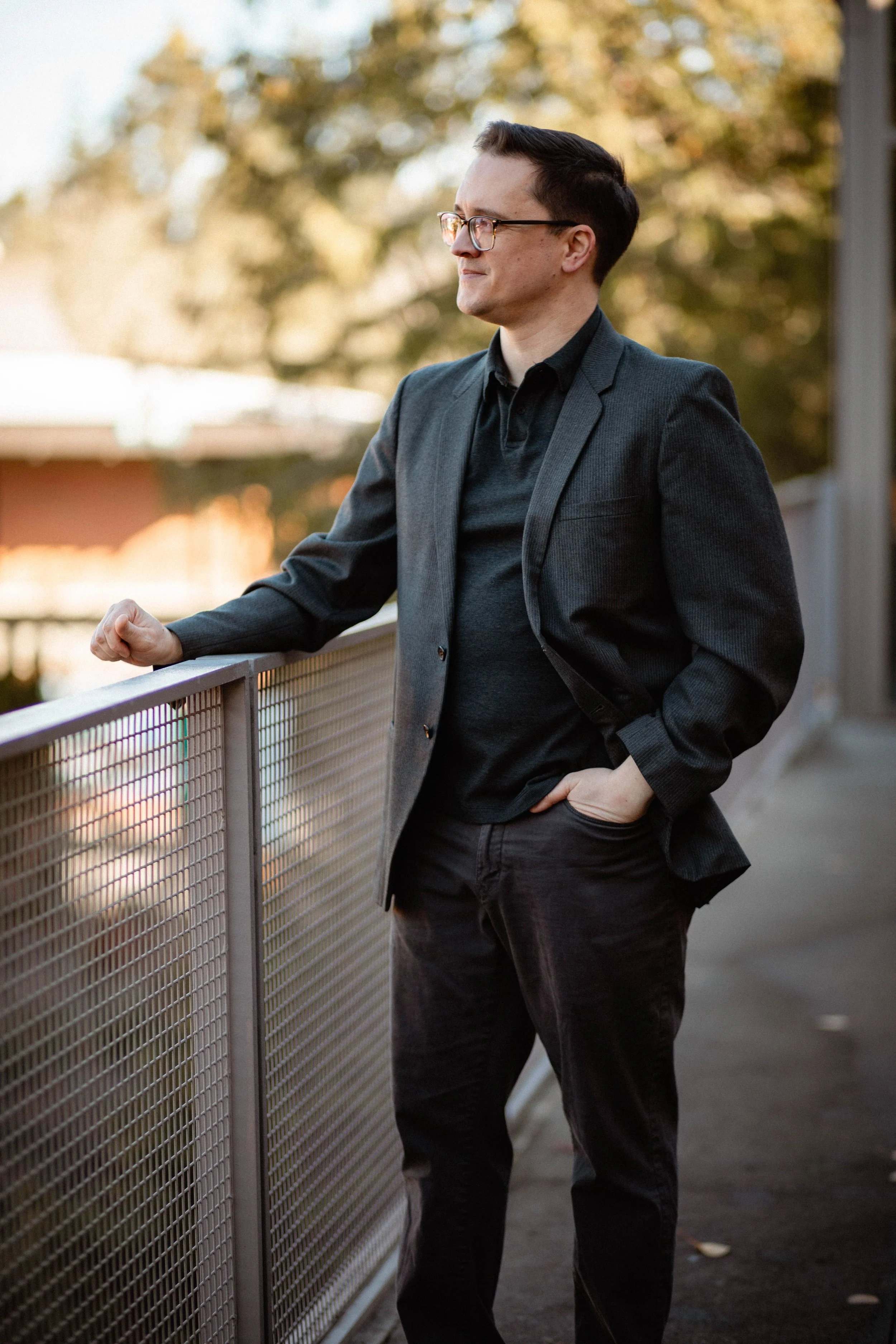 A man wearing glasses, a dark blazer, and dark pants, standing outdoors next to a metal railing, looking to the side with a slight smile, during the daytime with trees and a building in the background. Seattle professional head shot photography