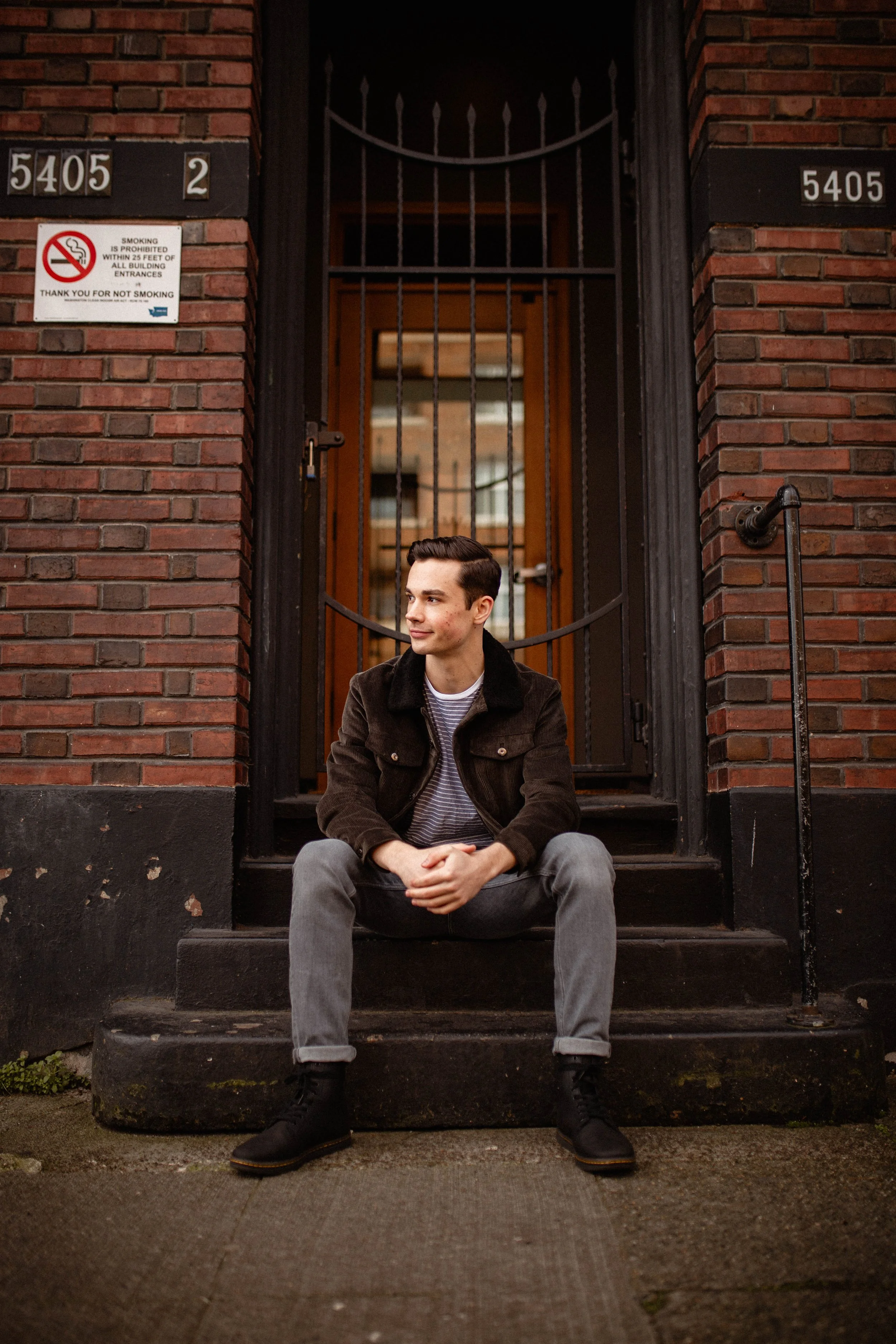Young man sitting on steps outside brick building, looking to the side, wearing a dark jacket, striped shirt, gray jeans, and black boots. Seattle professional head shot photography