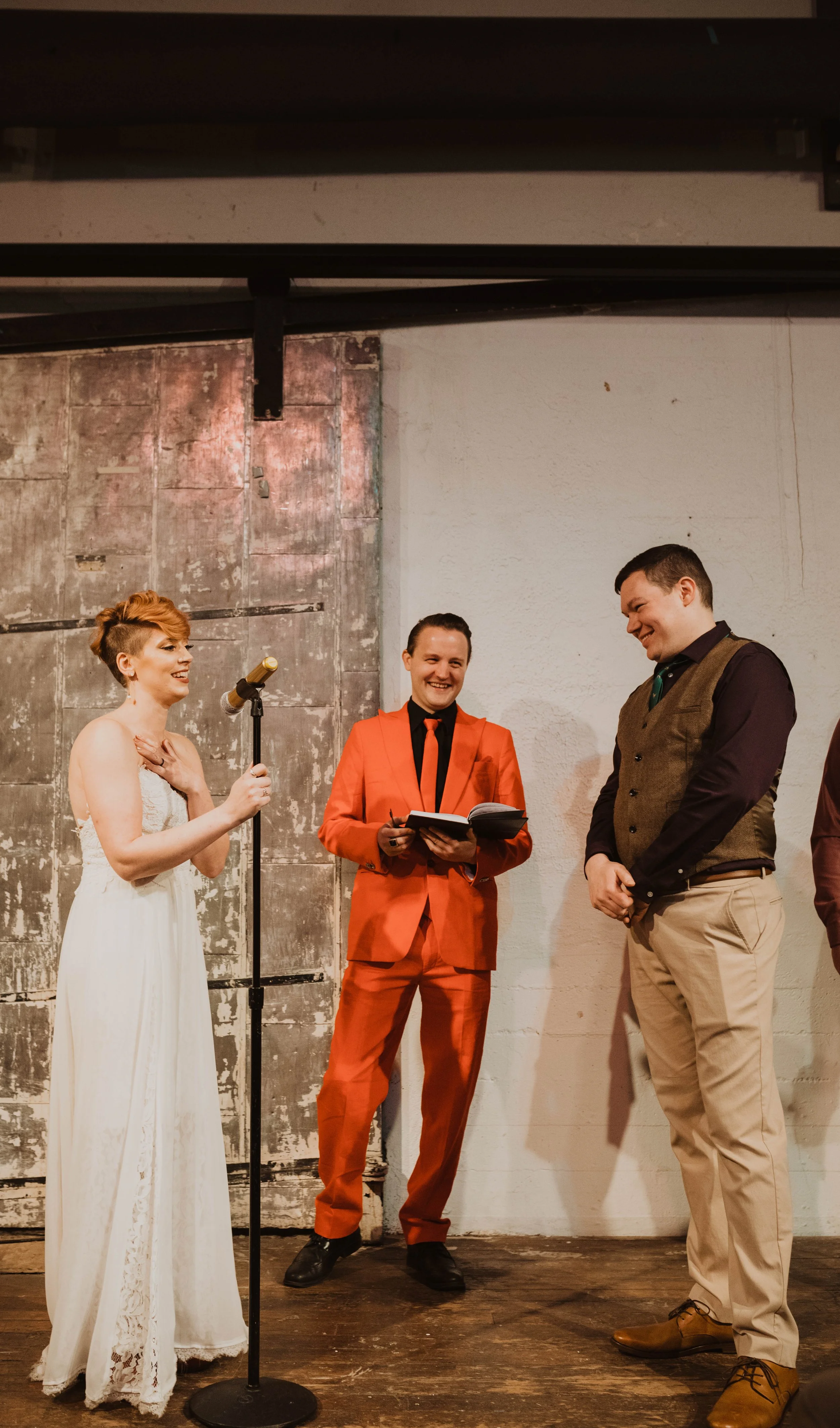A woman in a white dress appears to be getting married, standing in front of a mic stand, with two men in formal attire smiling and laughing during a wedding ceremony or celebration. Pioneer Square, Seattle, WA wedding photography.