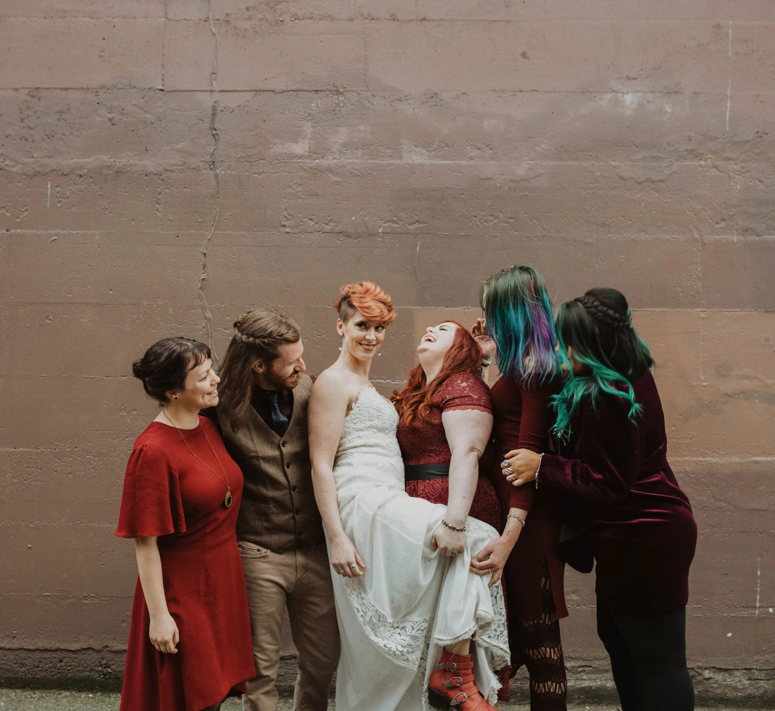 Group of six friends standing together in front of a plain brown wall, enjoying a moment of laughter, with one woman wearing a wedding dress. Pioneer Square, Seattle, WA wedding photography.