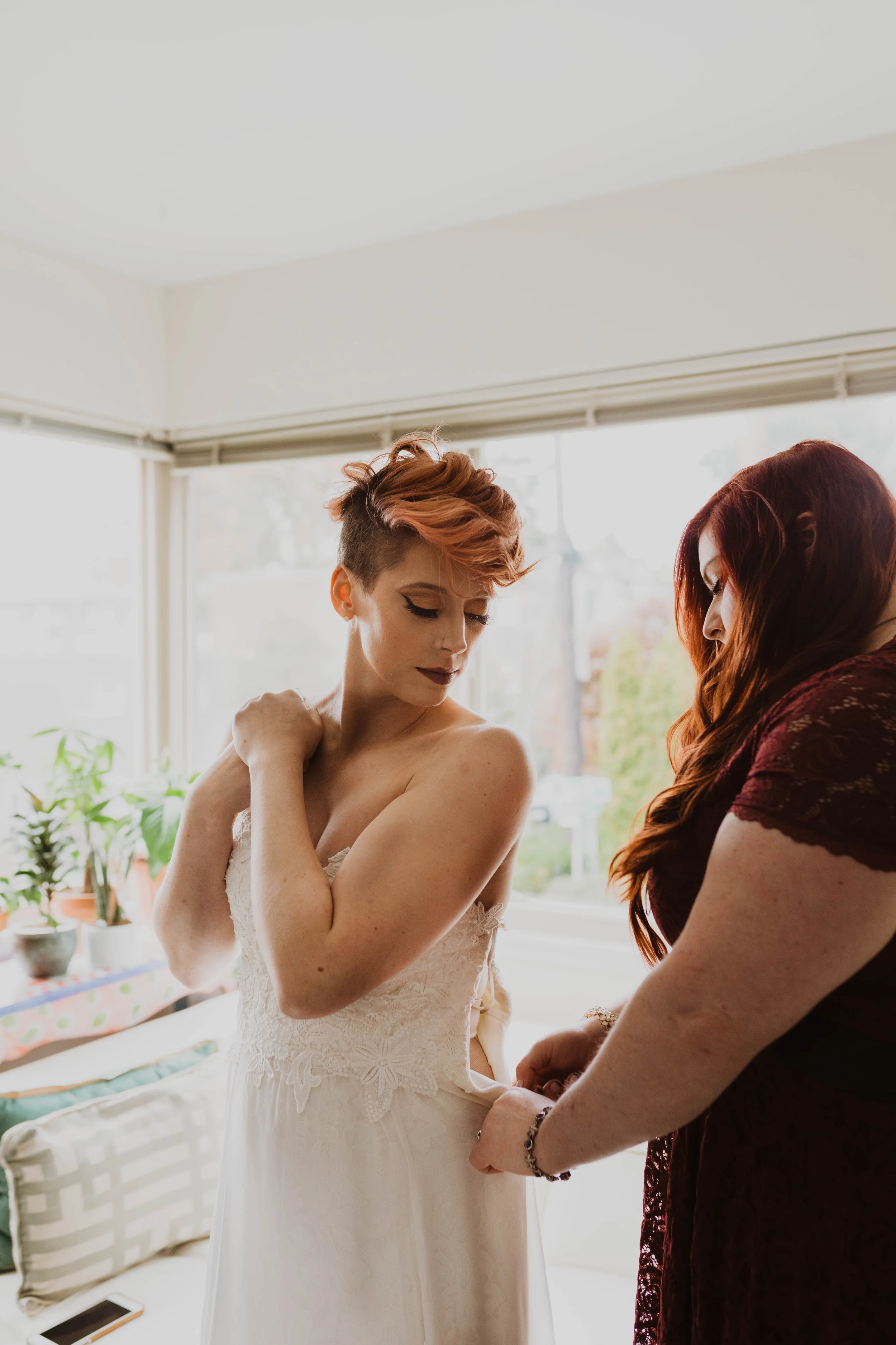 A bride in a white lace wedding dress having her dress adjusted by a woman in a burgundy dress inside a bright room with large windows and potted plants. Pioneer Square, Seattle, WA wedding photography. 