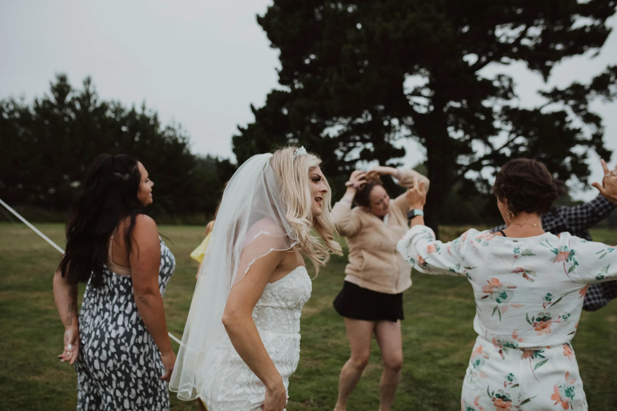 Group of women dancing outdoors on a grassy field during daytime, one woman dressed in a wedding gown and veil, others in casual summer clothes, celebrating at a wedding reception or outdoor event. Long Beach, WA wedding photography.