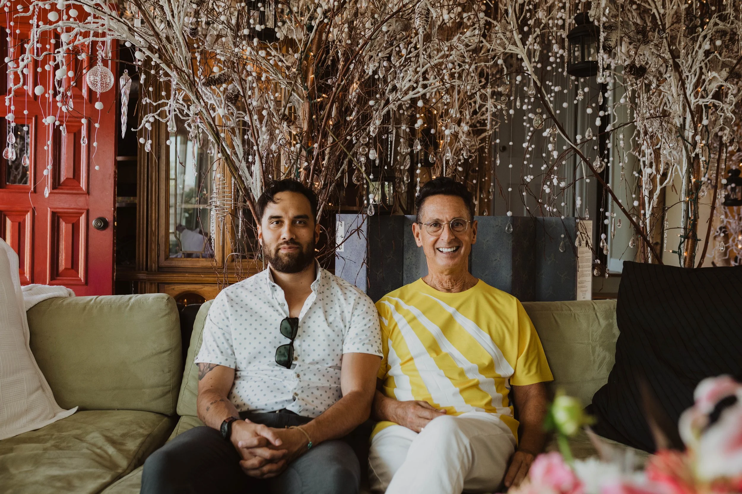 Two men sitting on a sofa in a decorated indoor setting with branches, hanging ornaments, and lights behind them. The man on the left has dark hair, a beard, and is wearing a white shirt with small patterns, sunglasses hanging on his shirt, and tatto
