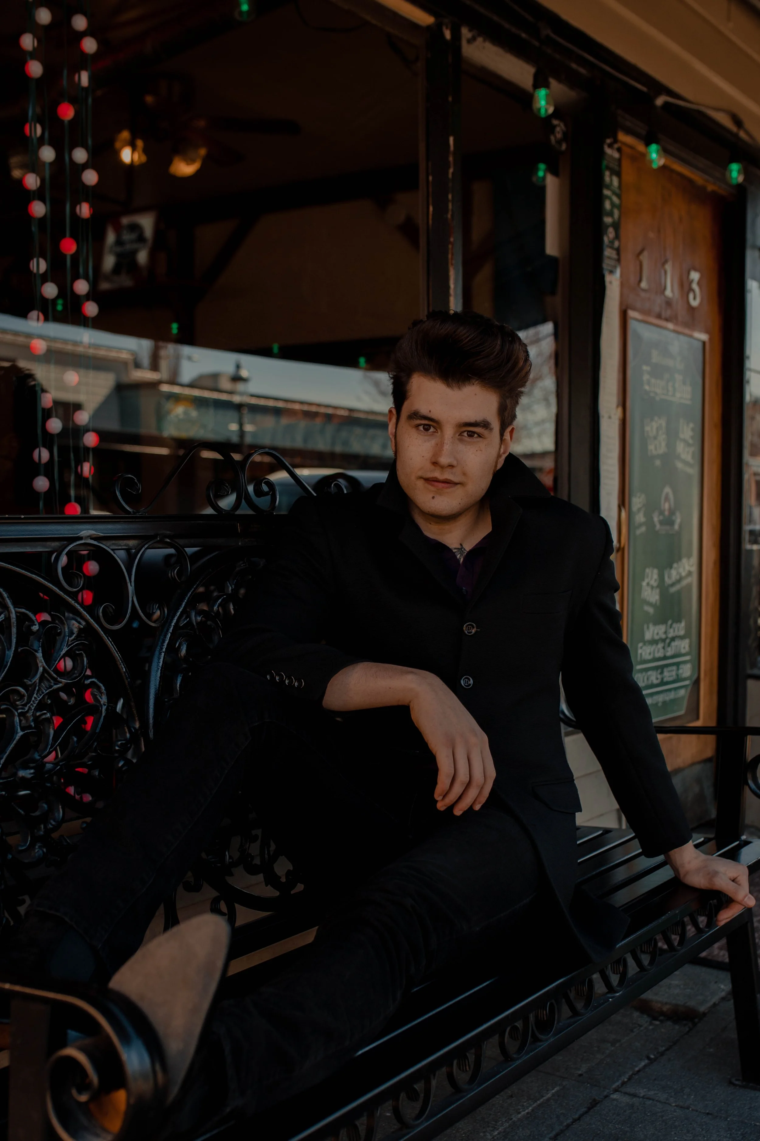 A young man in a black jacket sitting on a decorative black iron bench outside a restaurant or café. Seattle professional head shot photography