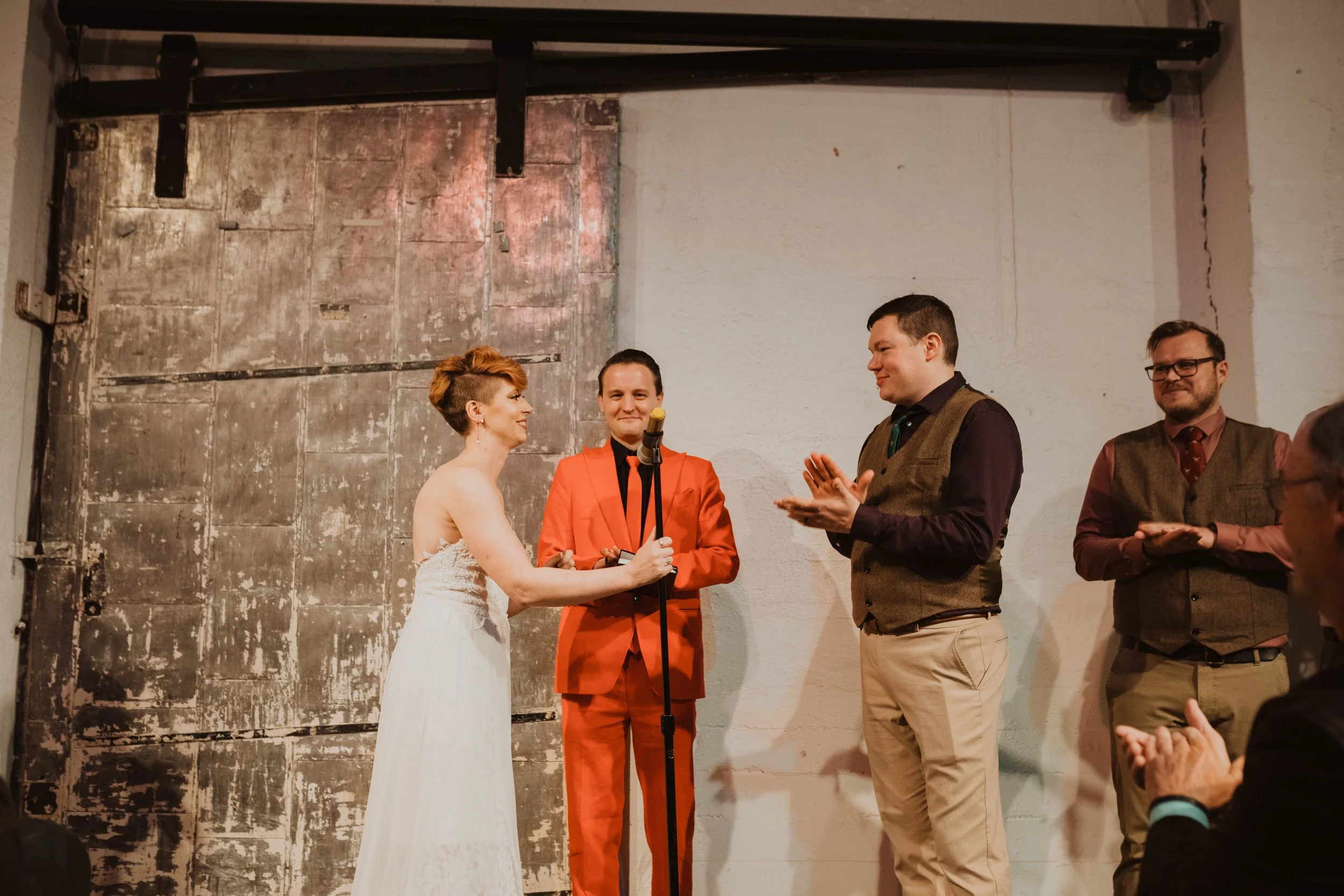 A wedding ceremony with a woman in a white dress holding a microphone and a man in a vest clapping, standing on a rustic stage with a distressed metal door in the background. Pioneer Square, Seattle, WA wedding photography.
