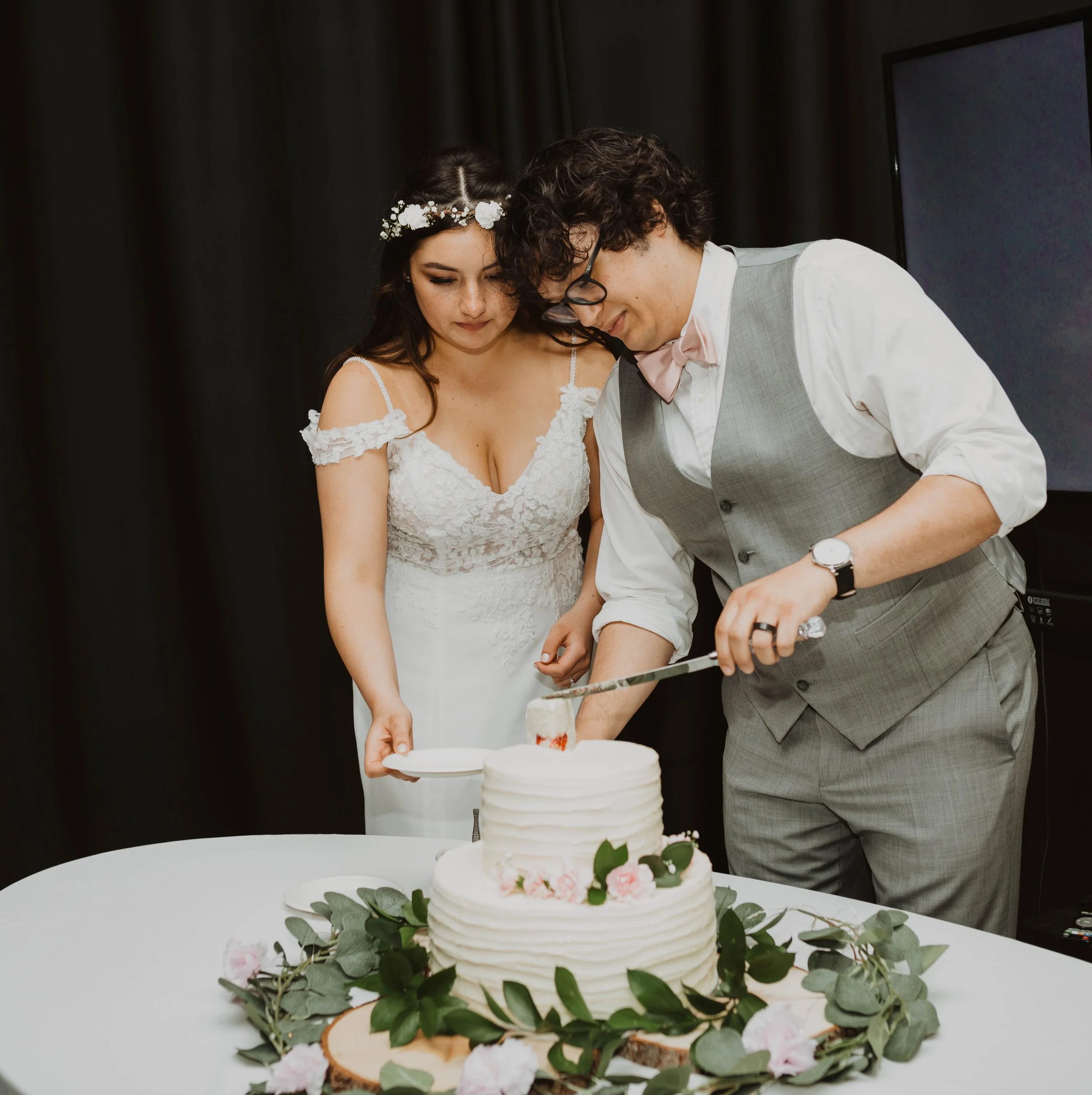 A bride in a white wedding dress and a man in a gray vest and pink bowtie cutting a wedding cake together at a celebration. Seattle, WA wedding photography.