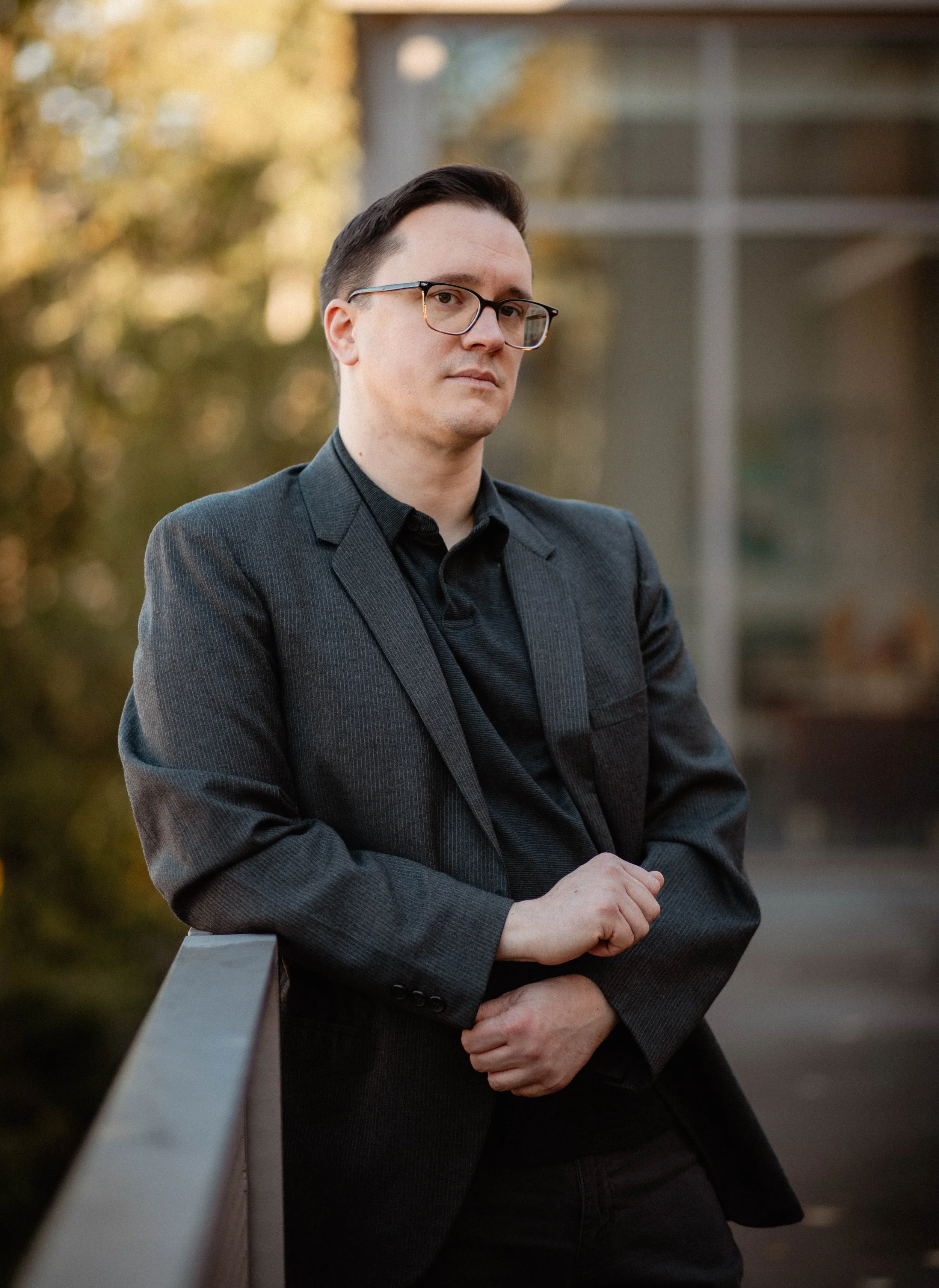 A man in a dark suit and glasses standing outdoors near a railing with a blurred background of trees and a modern building. Seattle professional head shot photography