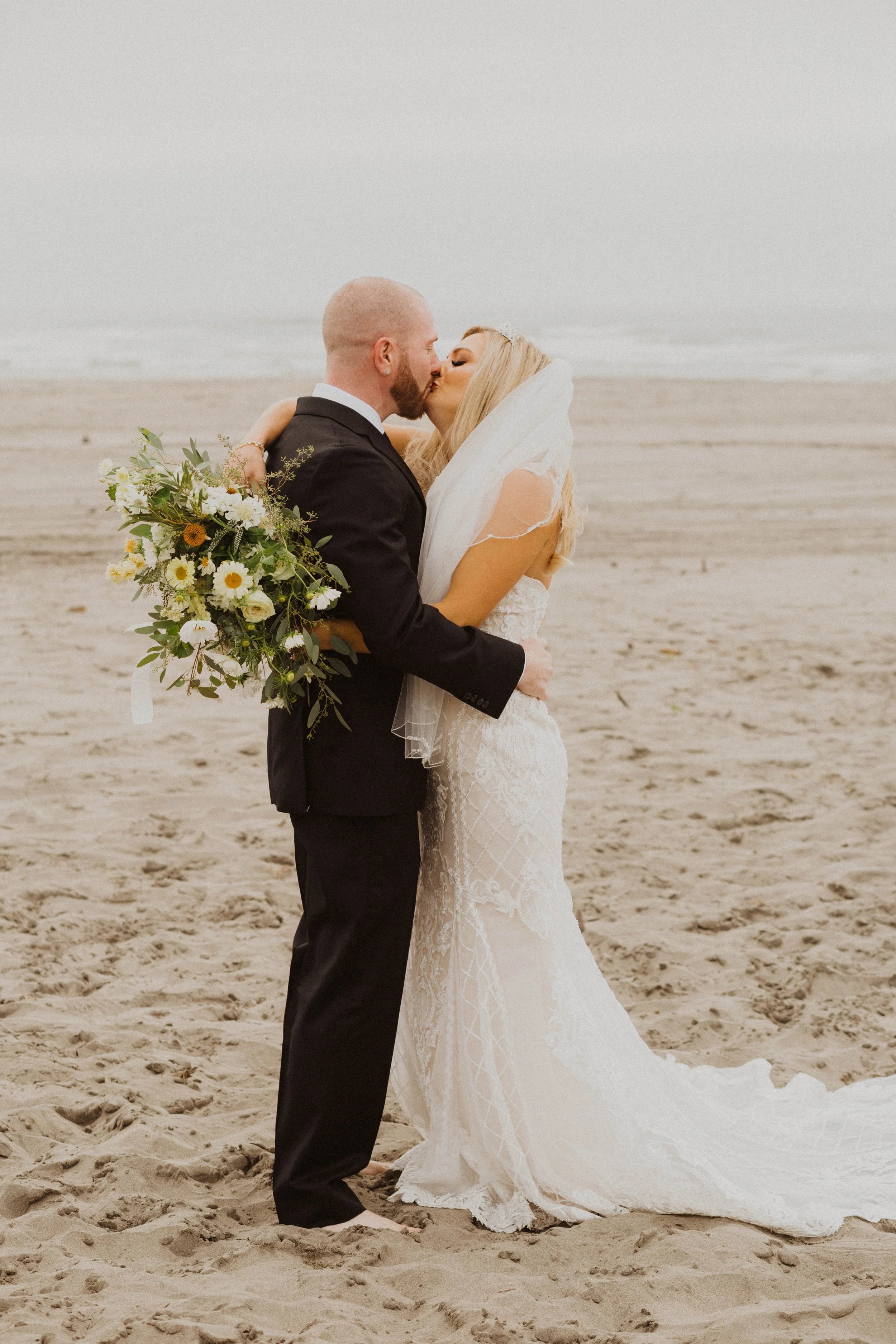 A bride and groom kissing on the beach, with the bride holding a bouquet, during their wedding. Long Beach, WA wedding photography.